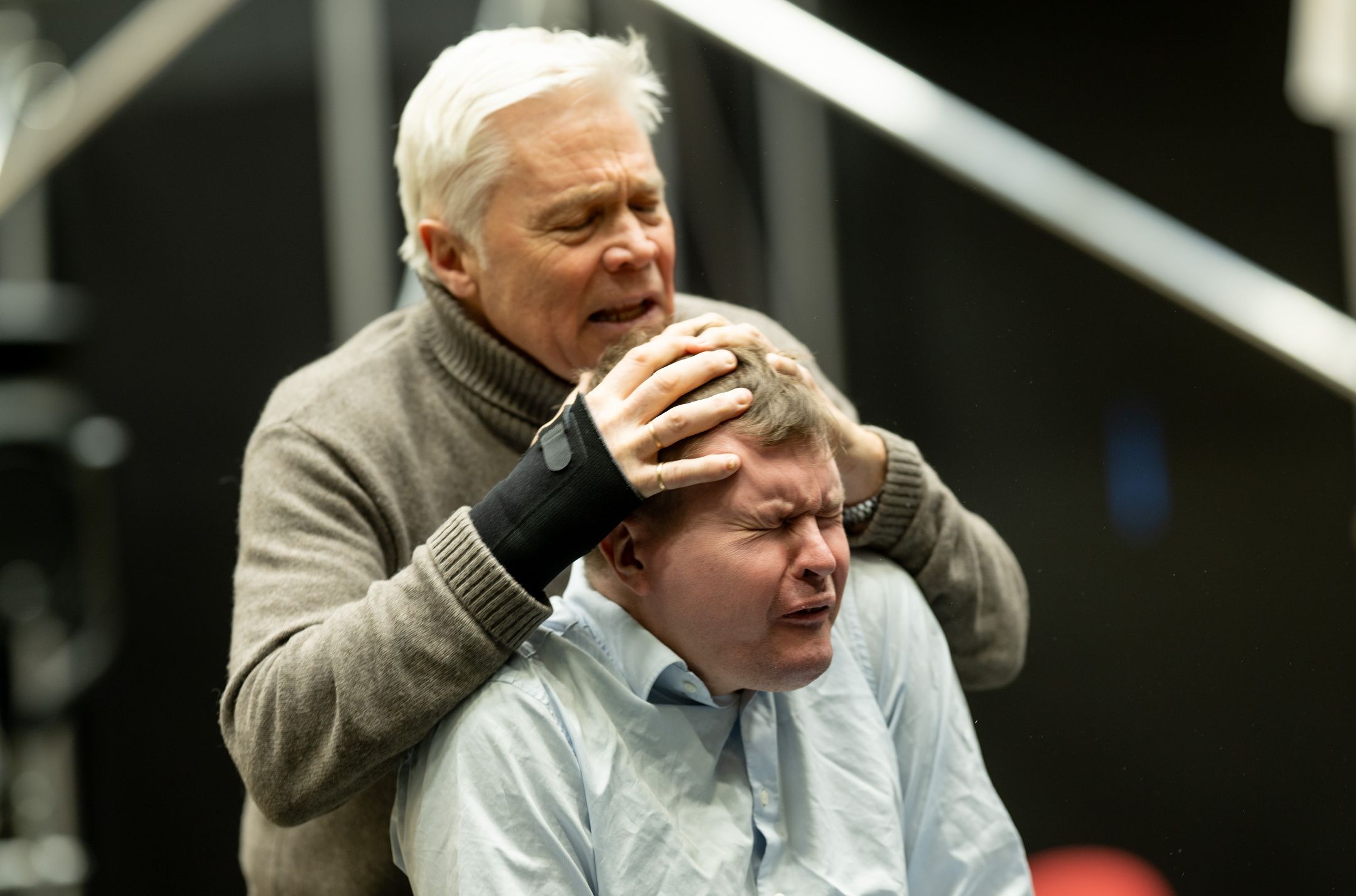 En mann holder hodet til en annen mann med lukkede øyne og smertefullt uttrykk under en intens sceneprøve.