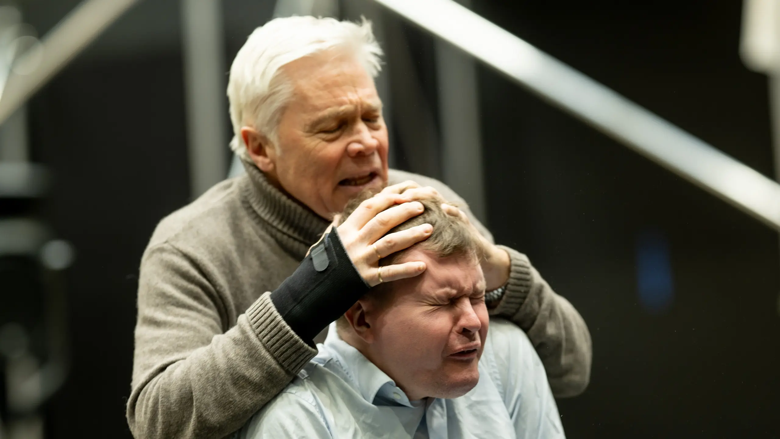 En mann holder hodet til en annen mann med lukkede øyne og smertefullt uttrykk under en intens sceneprøve.