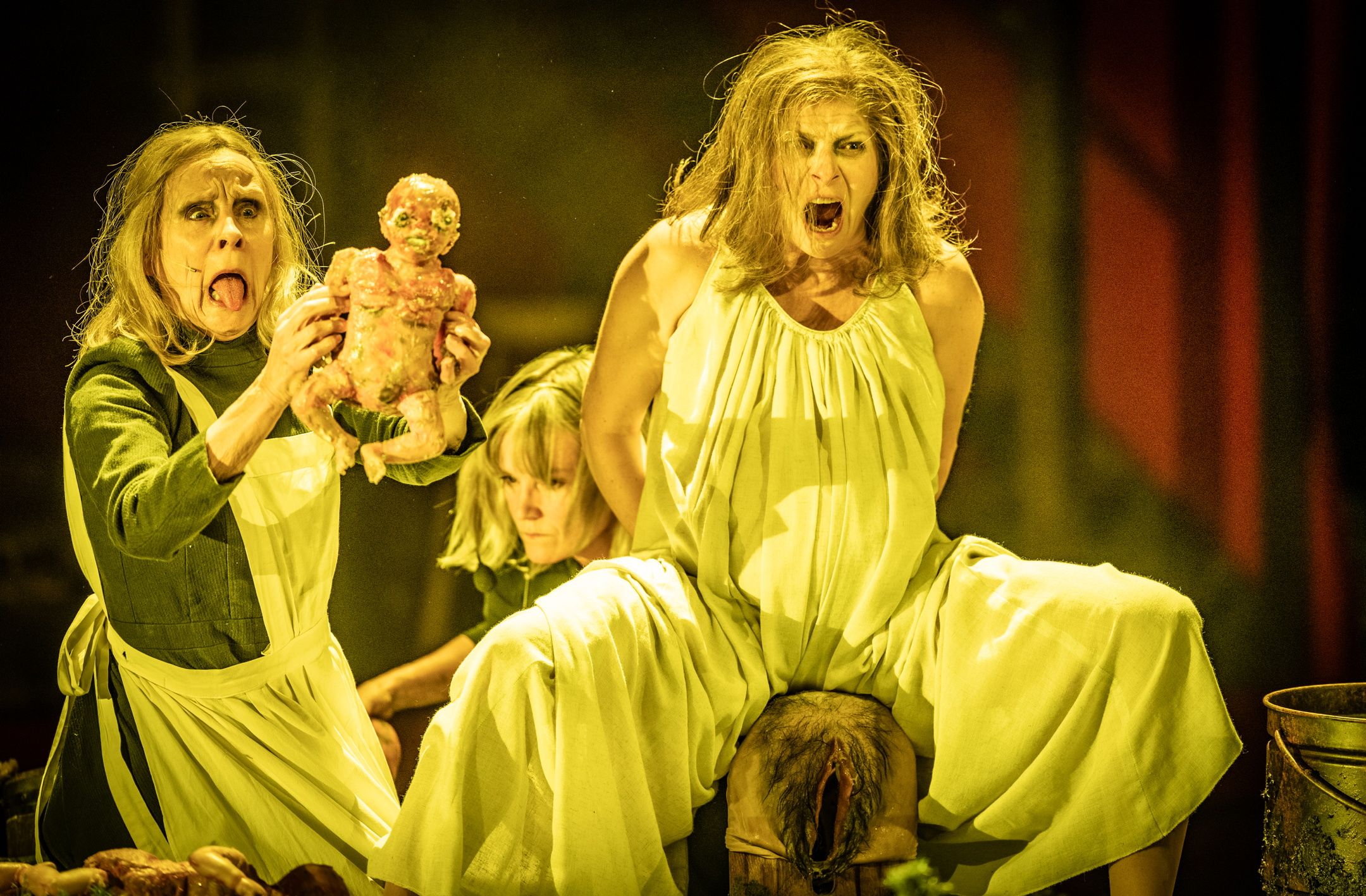To kvinner i gule kjoler roper mens en holder opp en blodig dukke, i en scene med sterk fødselsmetafor og teatralsk uttrykk.