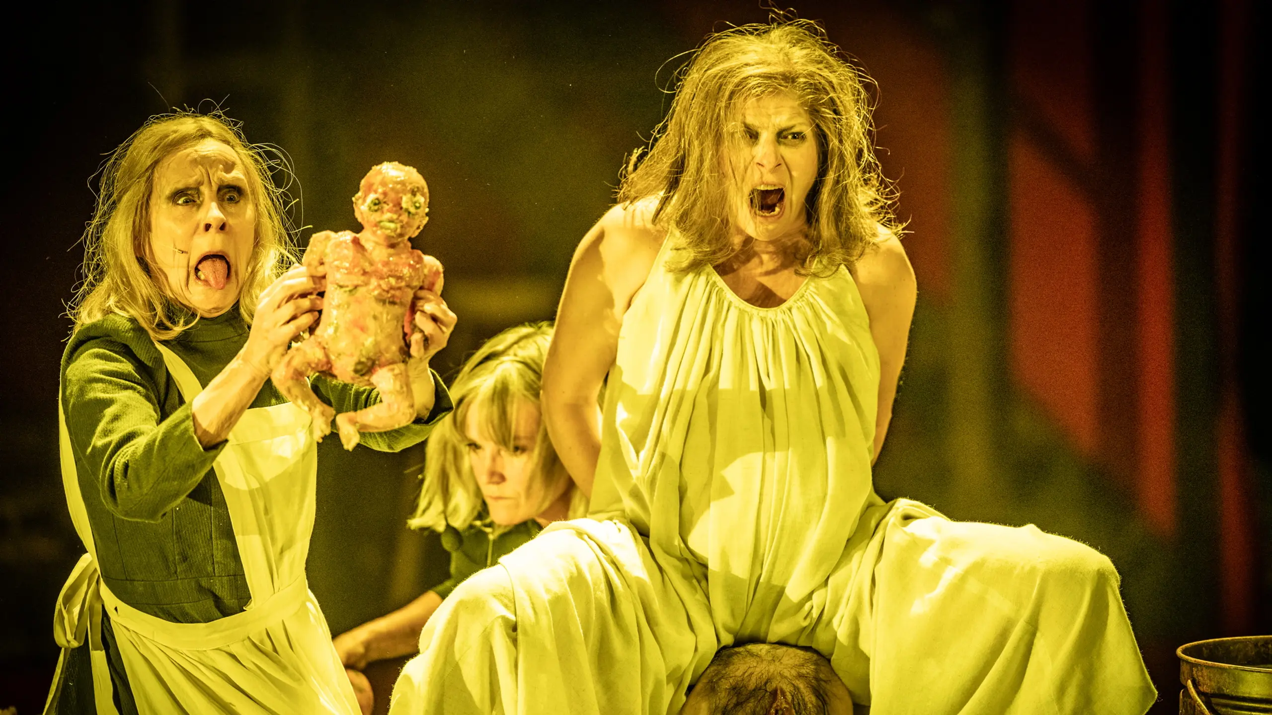 To kvinner i gule kjoler roper mens en holder opp en blodig dukke, i en scene med sterk fødselsmetafor og teatralsk uttrykk.