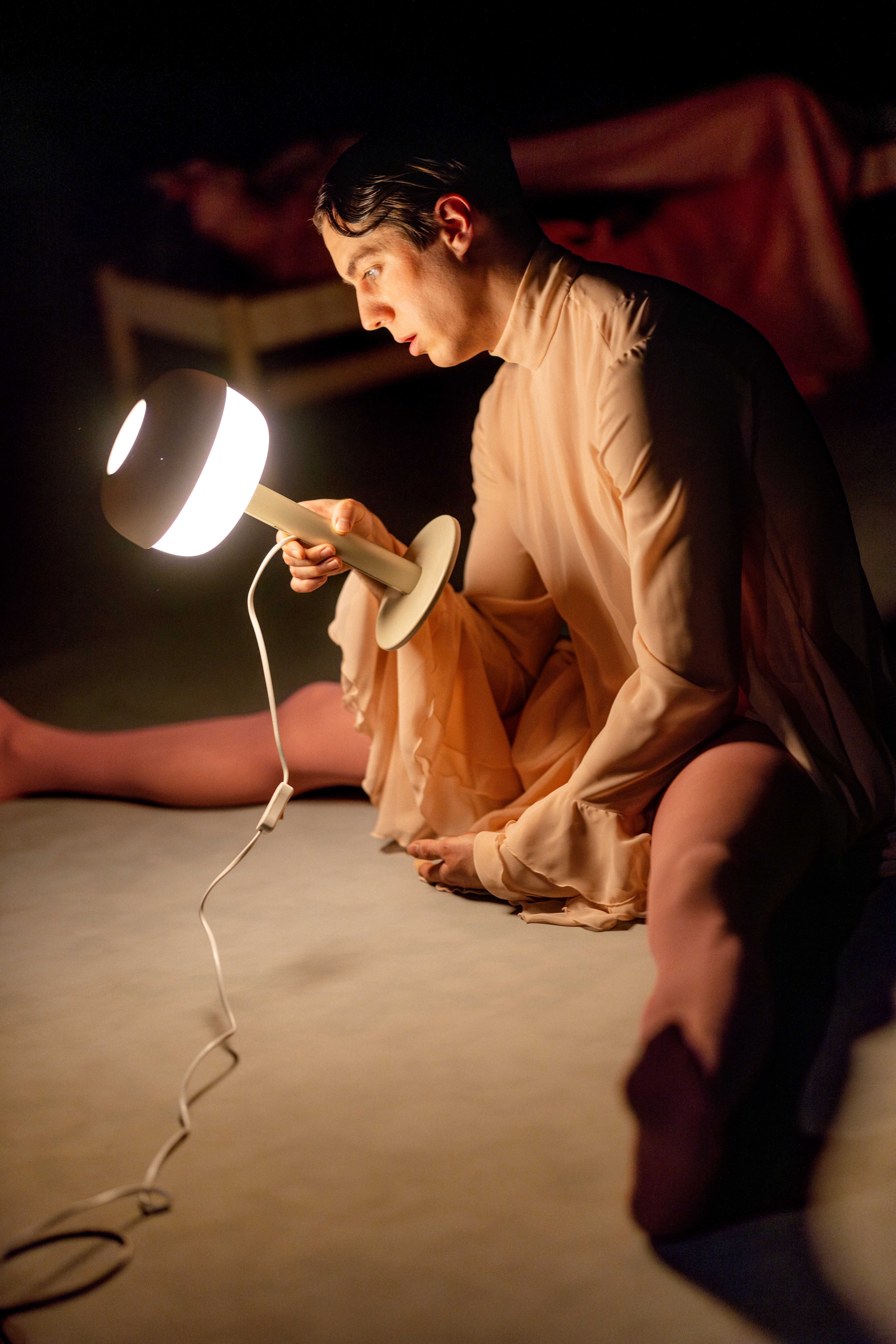 Person i lys kostyme sitter på gulvet og holder en bordlampe som lyser opp ansiktet i mørket