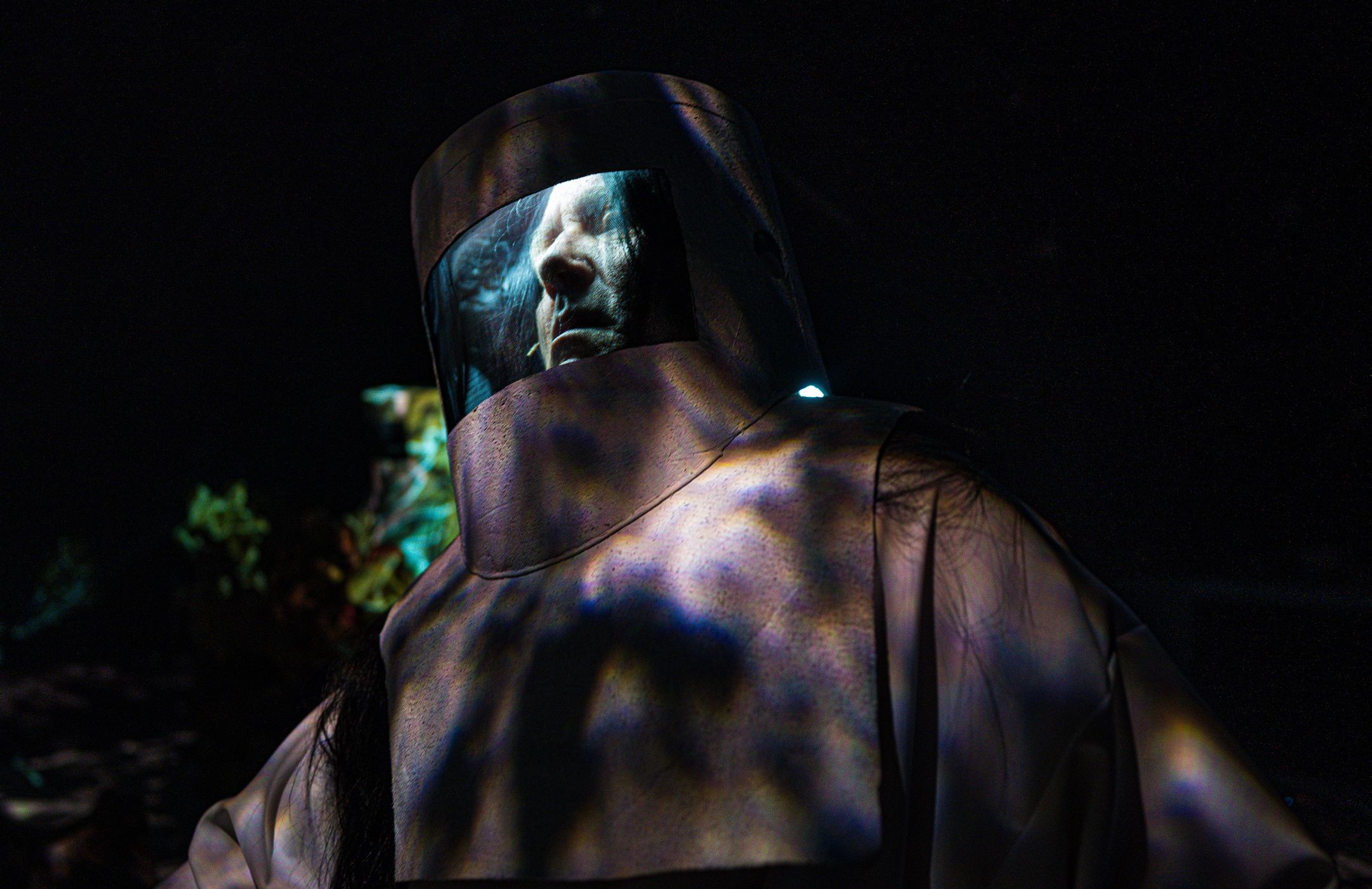 Person i mørkt skogmiljø iført tung beskyttelsesdrakt med visirhjelm, ansikt synlig bak glasset