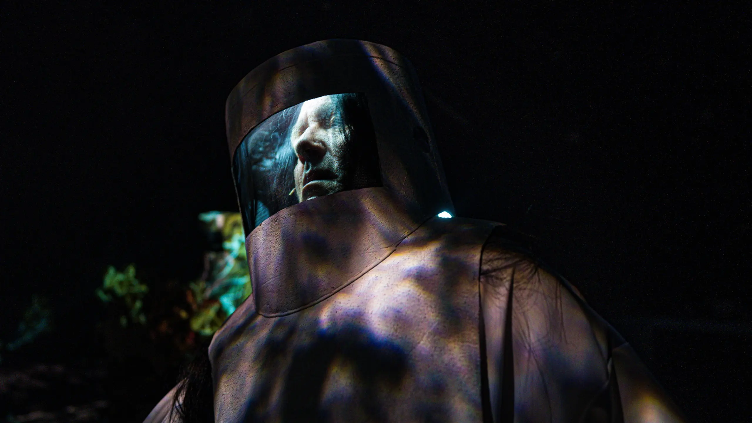 Person i mørkt skogmiljø iført tung beskyttelsesdrakt med visirhjelm, ansikt synlig bak glasset