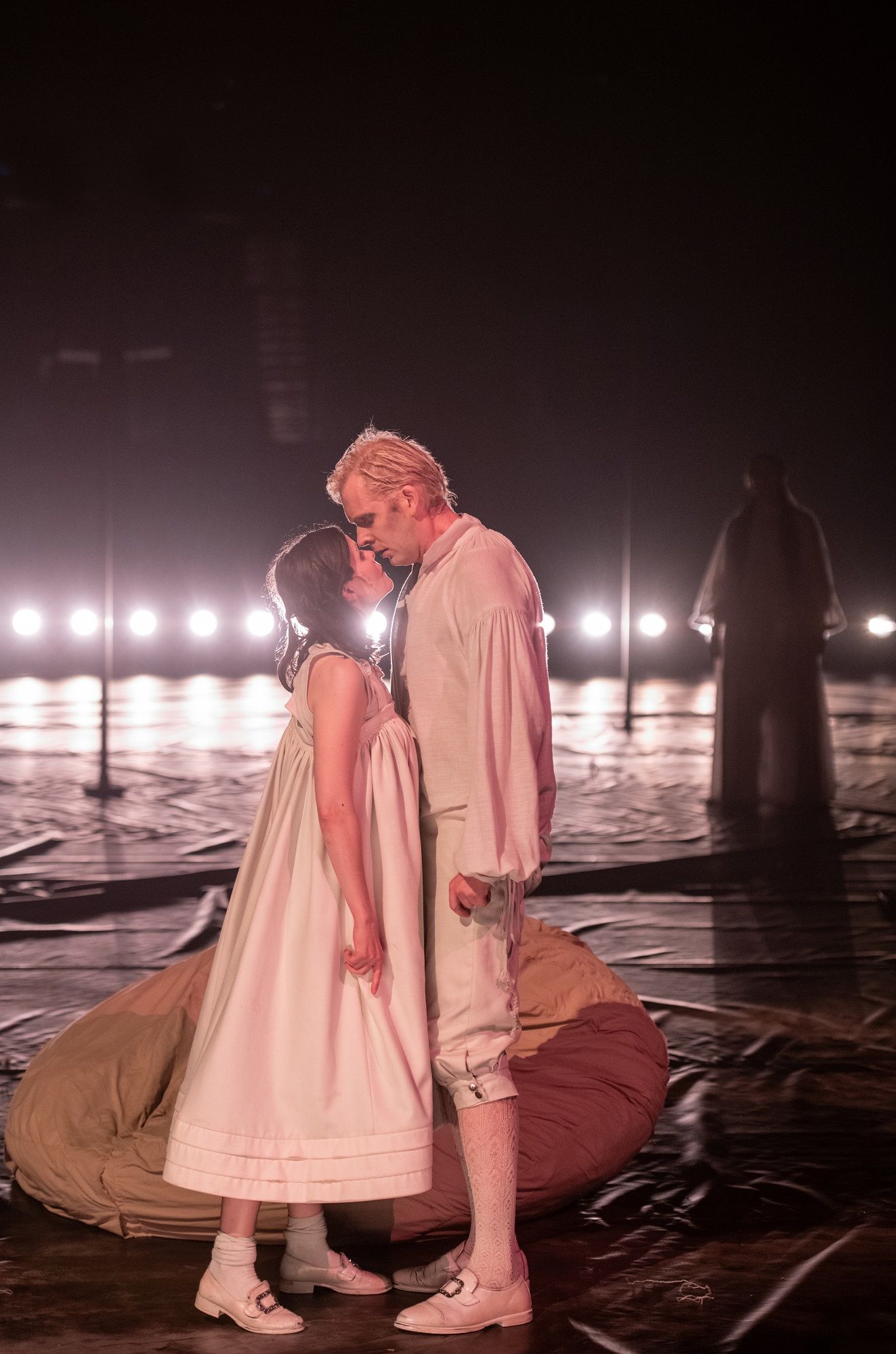 To skuespillere står tett sammen for å kysse iført hvite kostymer. I bakgrunnen en sanger.