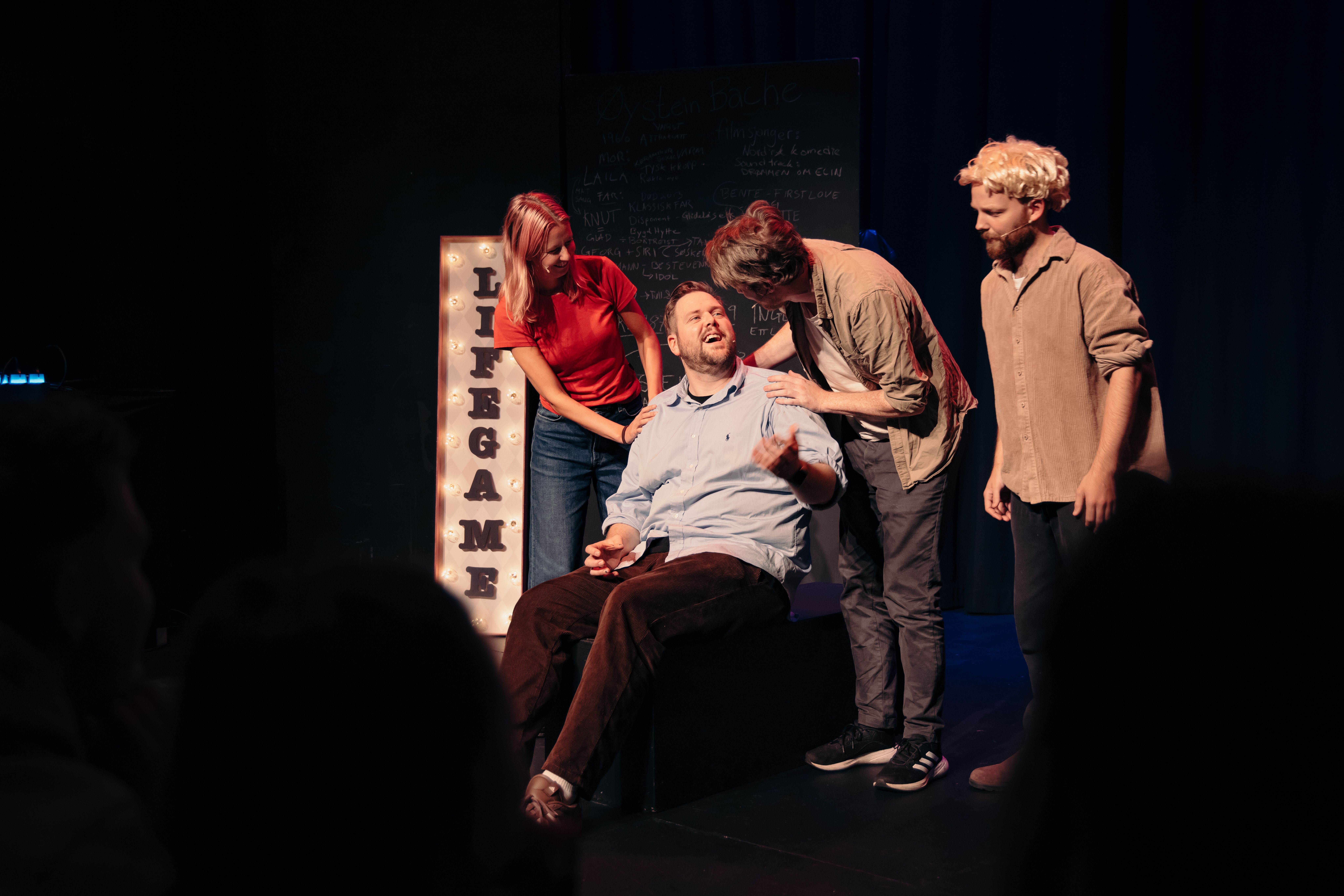 Flere personer rundt en sittende mann på scene, de lener seg inn og snakker mens skiltet LIFE GAME lyser bak