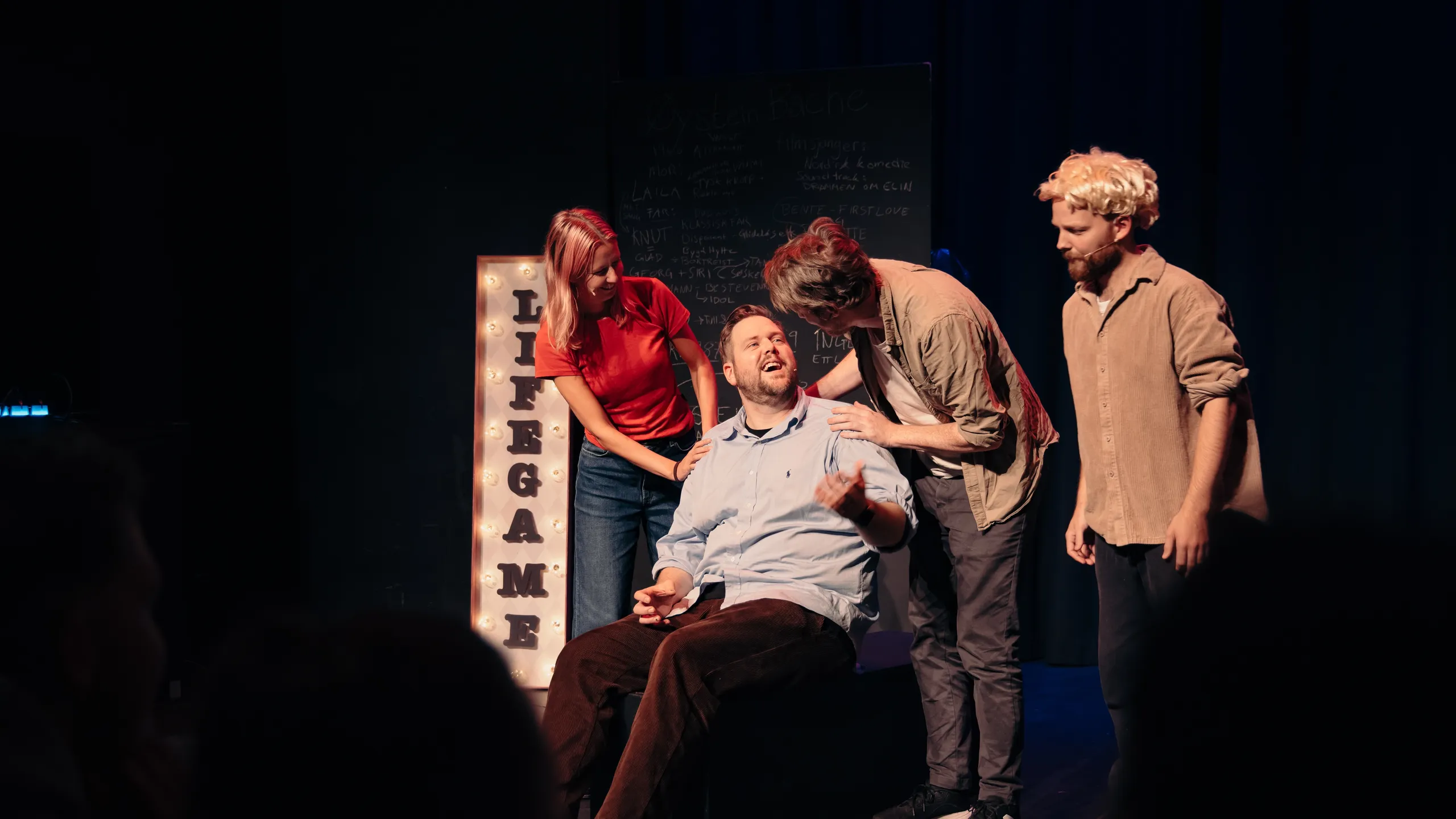 Flere personer rundt en sittende mann på scene, de lener seg inn og snakker mens skiltet LIFE GAME lyser bak