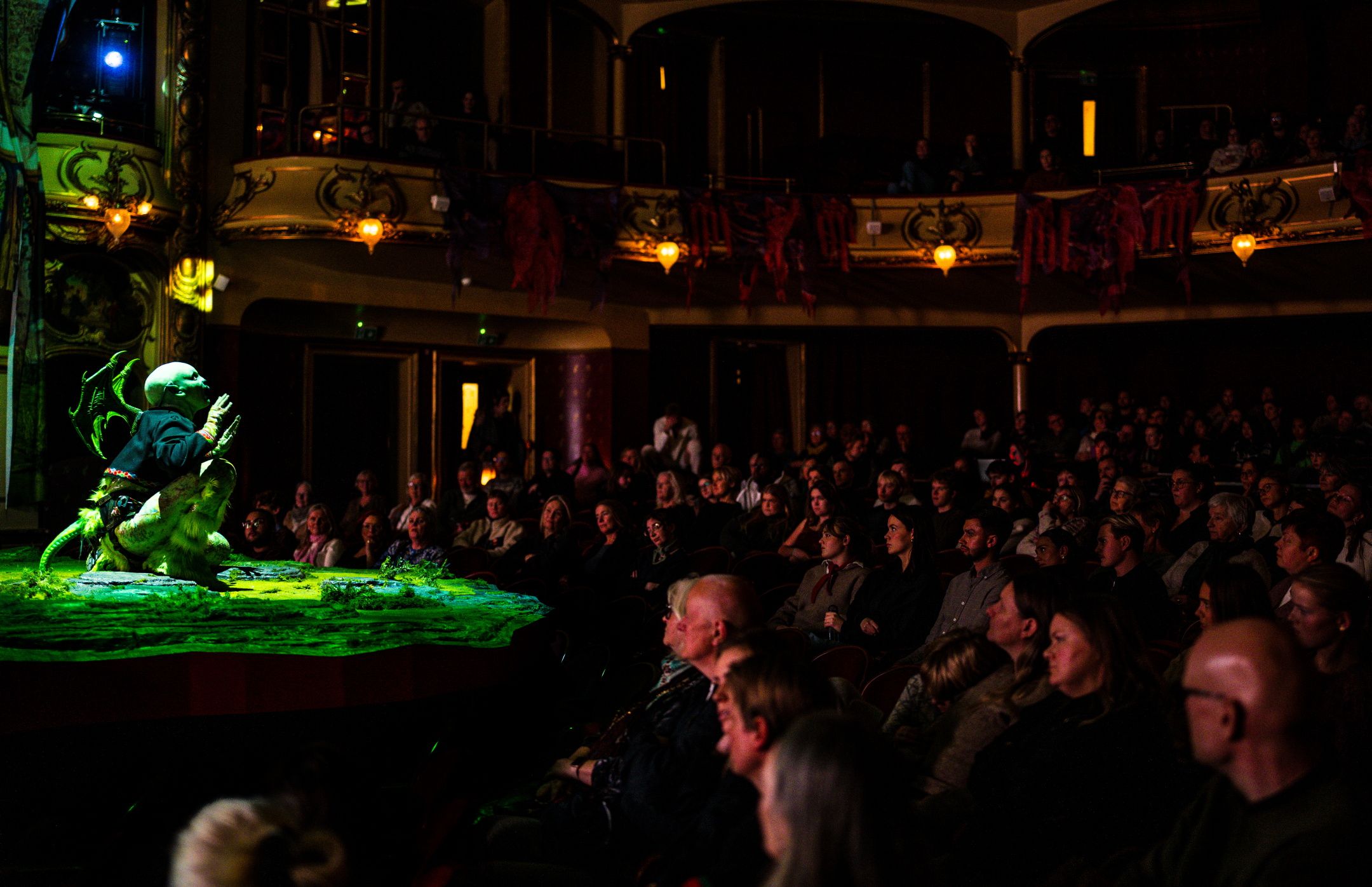 En publikumsal full av mennesker ser mot en liten scene der en skuespiller med dragevinger sitter i grønt scenelys