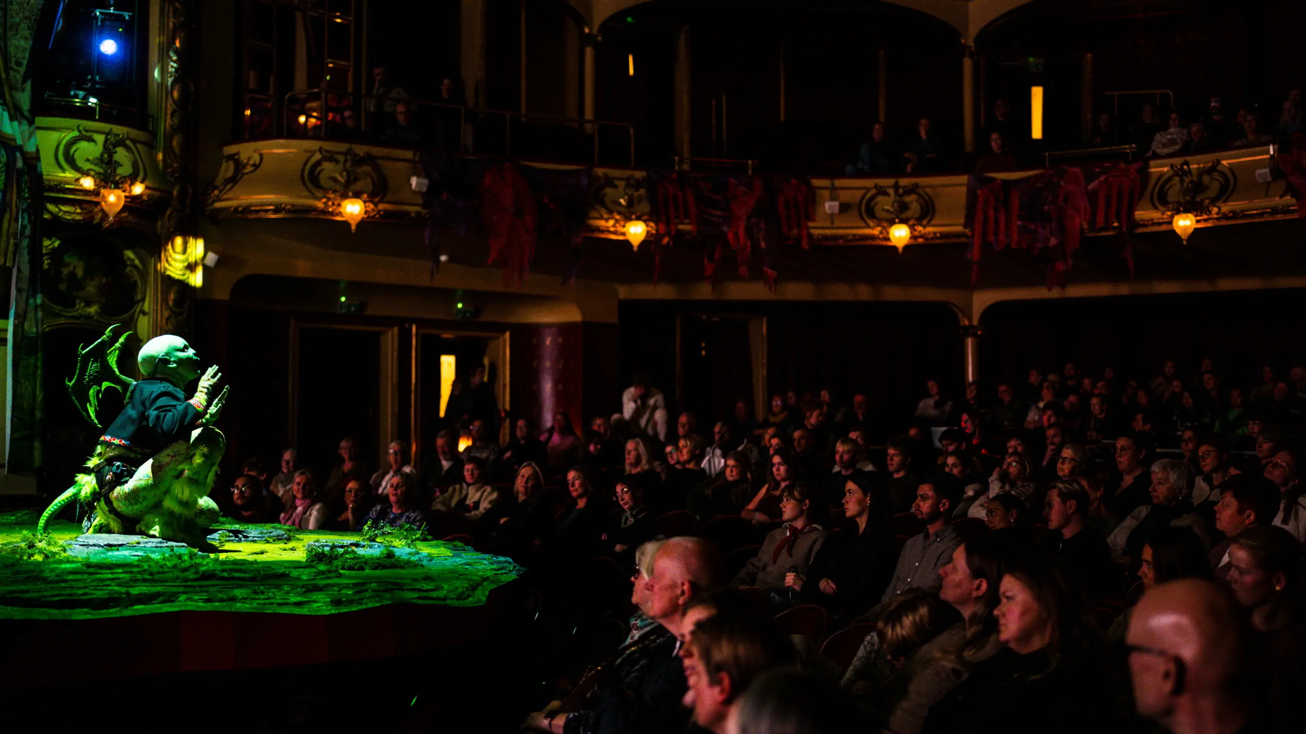 En publikumsal full av mennesker ser mot en liten scene der en skuespiller med dragevinger sitter i grønt scenelys
