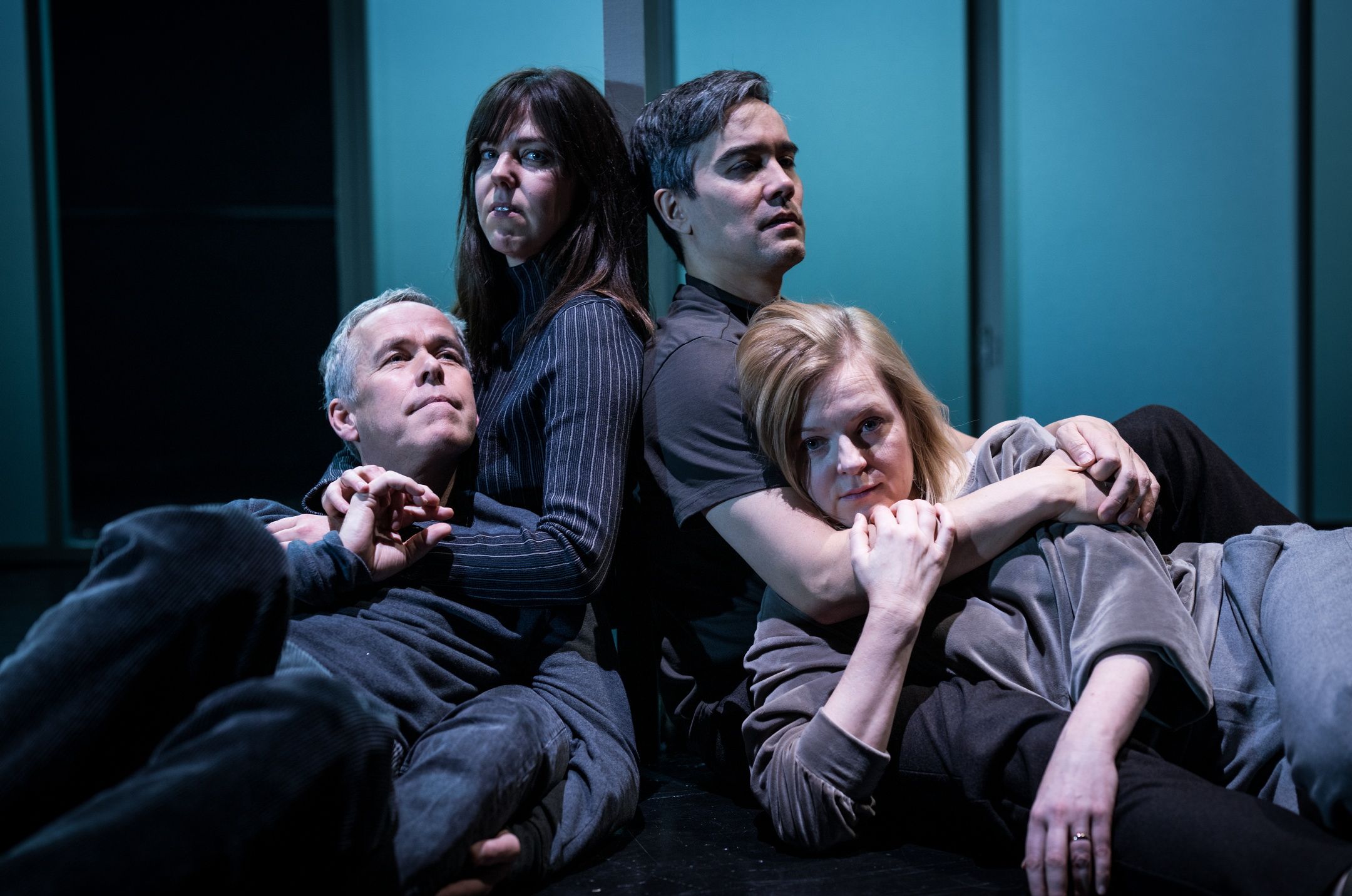 Fire voksne personer ligger på scenegulvet i en tett, oppstilt gruppe, med blågrønt lys bak dem og uttrykksfulle ansikter.