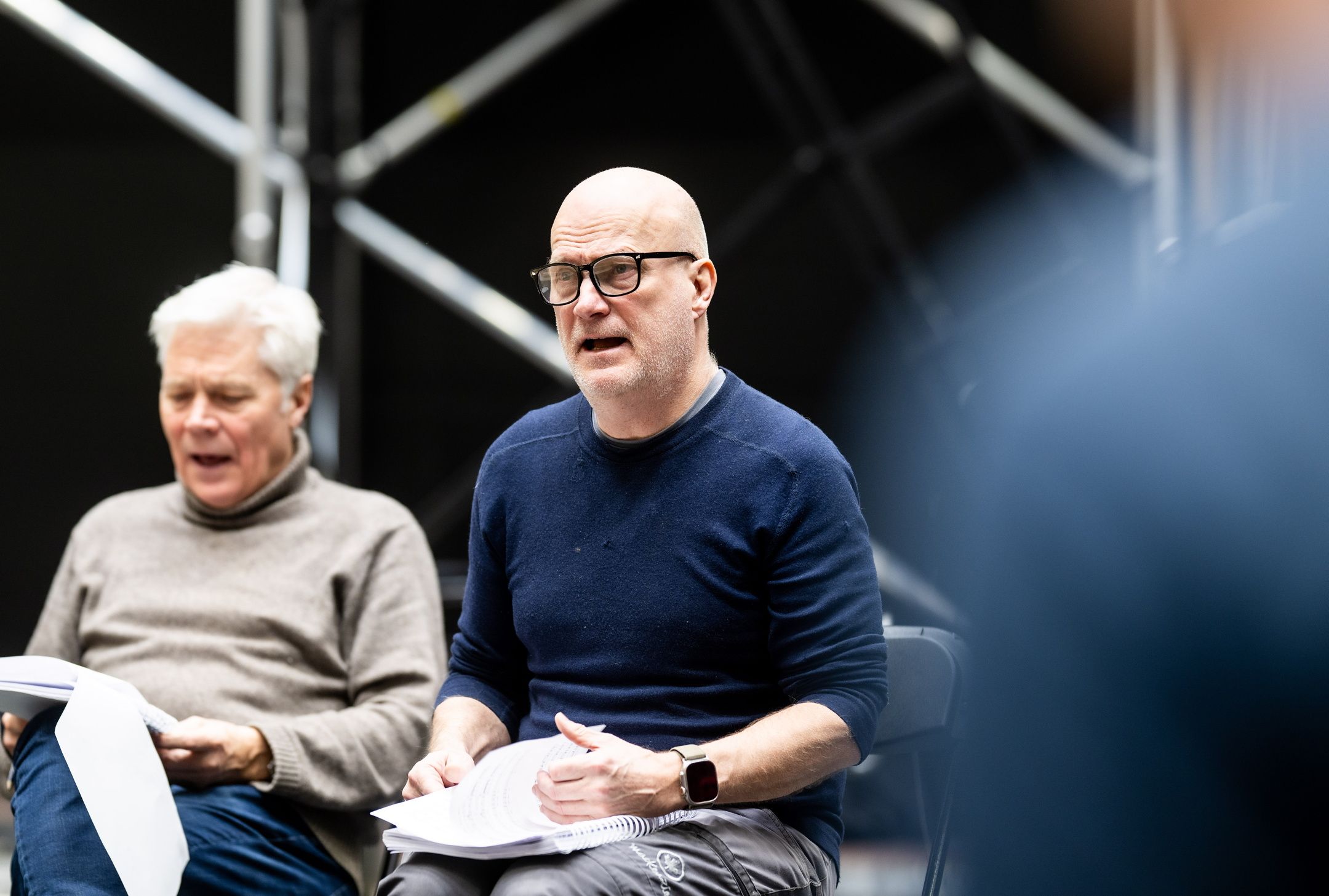 To menn leser manus under en bordprøve, med sceneteknisk rigg i bakgrunnen.