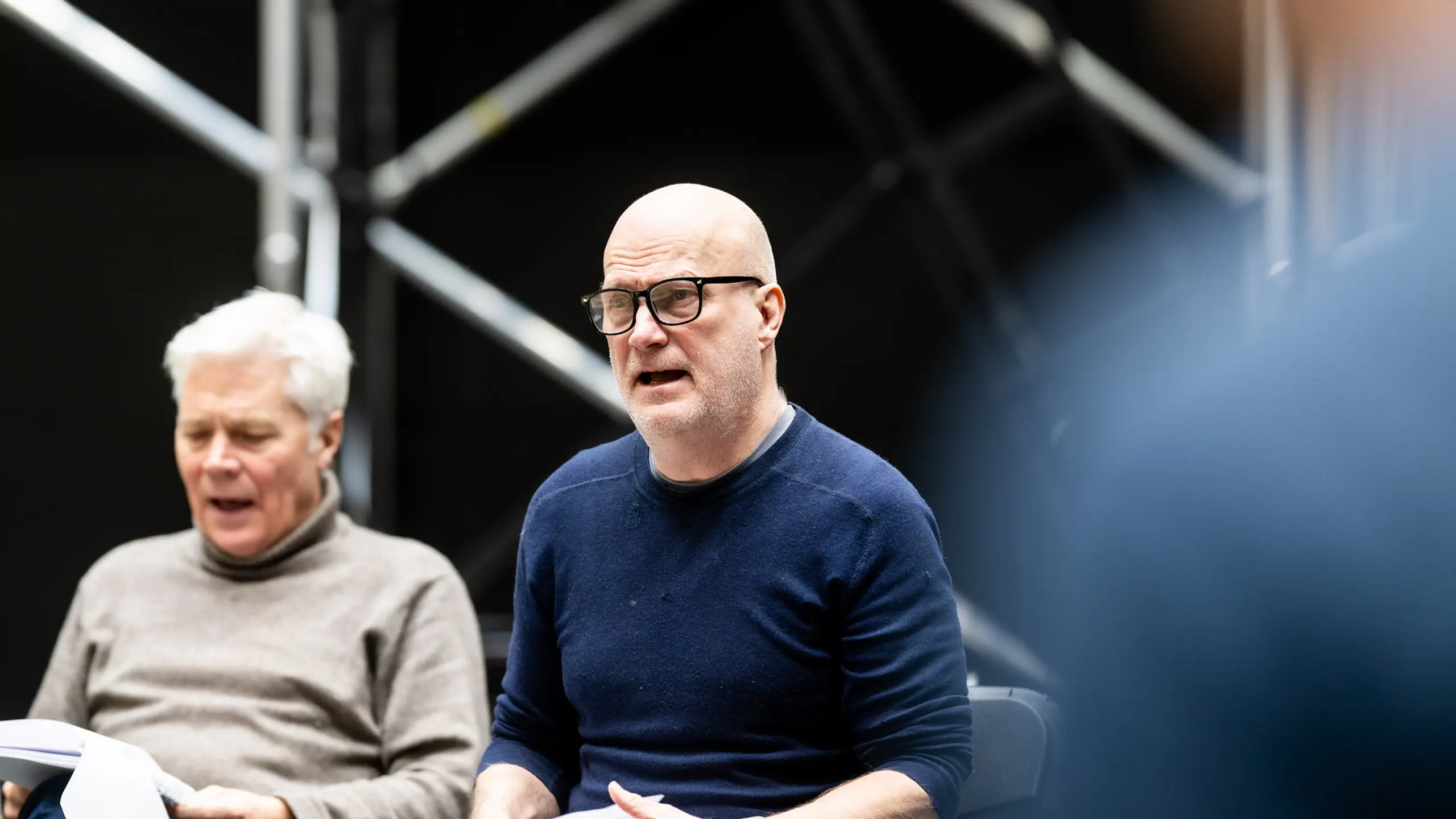 To menn leser manus under en bordprøve, med sceneteknisk rigg i bakgrunnen.