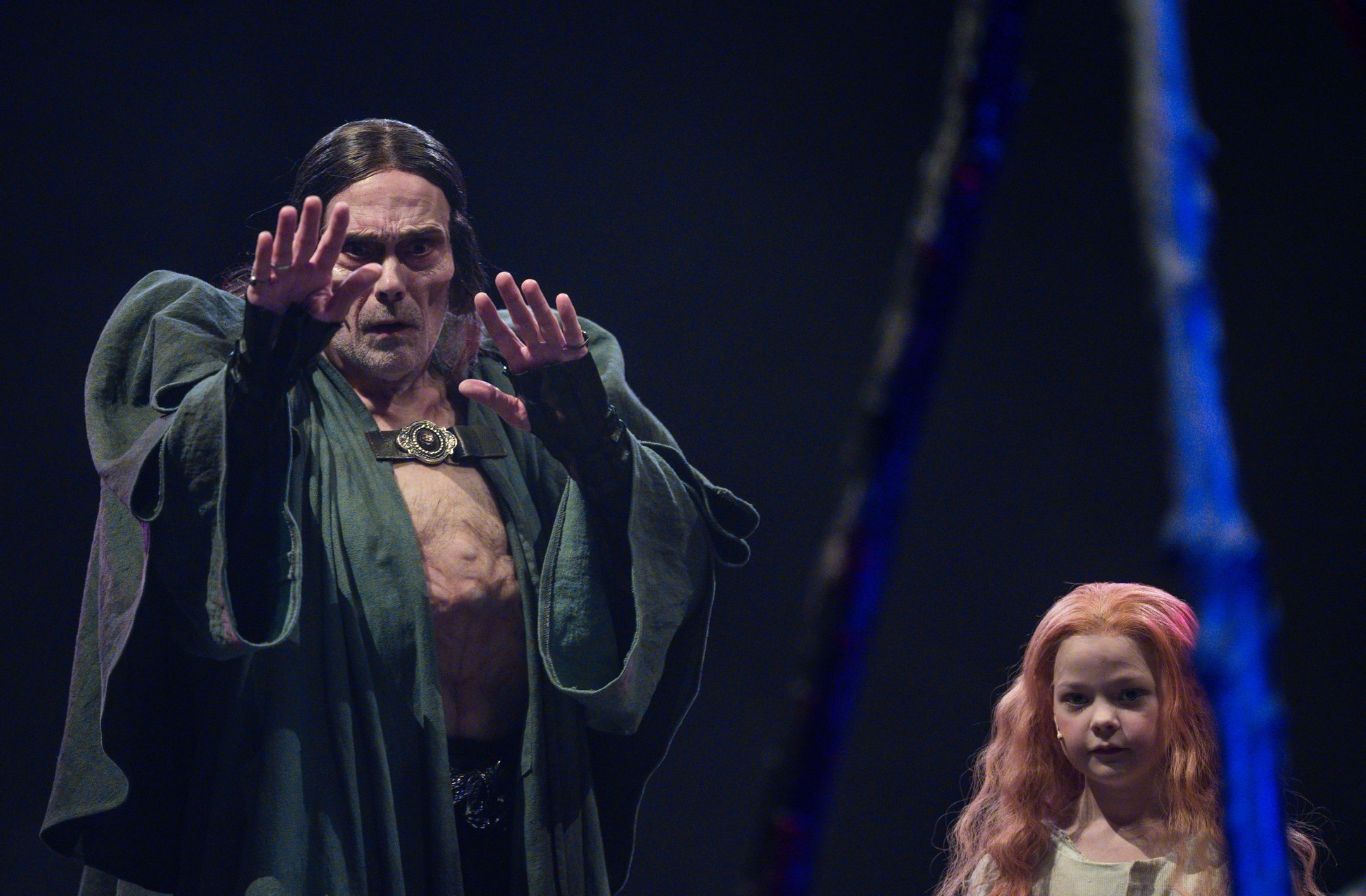 mannlig skuespiller med langt mørkt hår og åpen kappe strekker hendene fremover, mens en ung jente med langt rødt hår står rolig ved siden av ham.