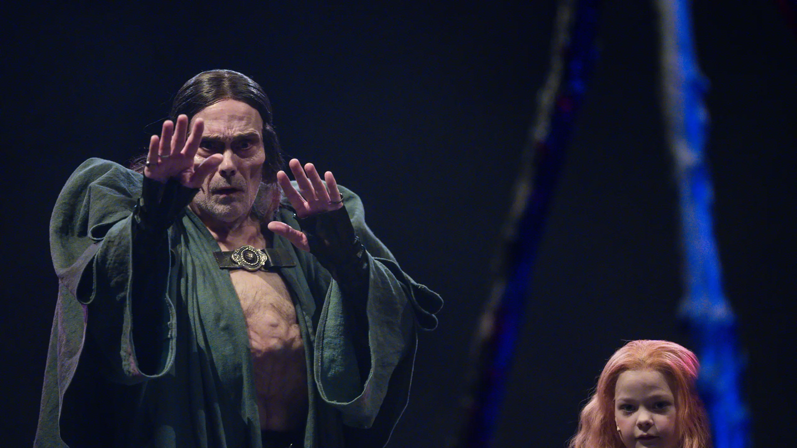 mannlig skuespiller med langt mørkt hår og åpen kappe strekker hendene fremover, mens en ung jente med langt rødt hår står rolig ved siden av ham.