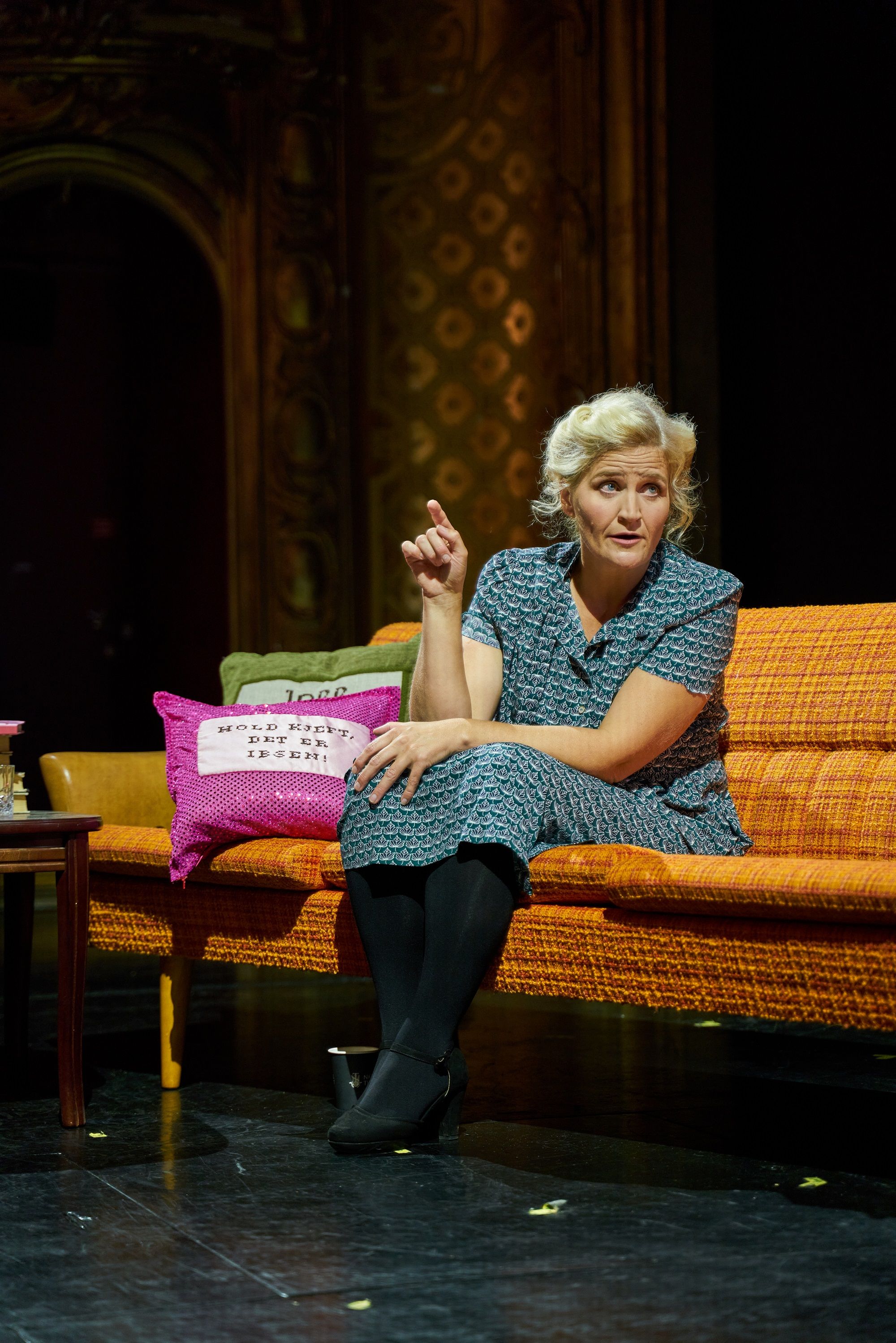 Kvinne sitter i en oransje sofa og gestikulerer, ved siden av henne ligger to puter med teksten "Hold kjeft, det er Ibsen!"