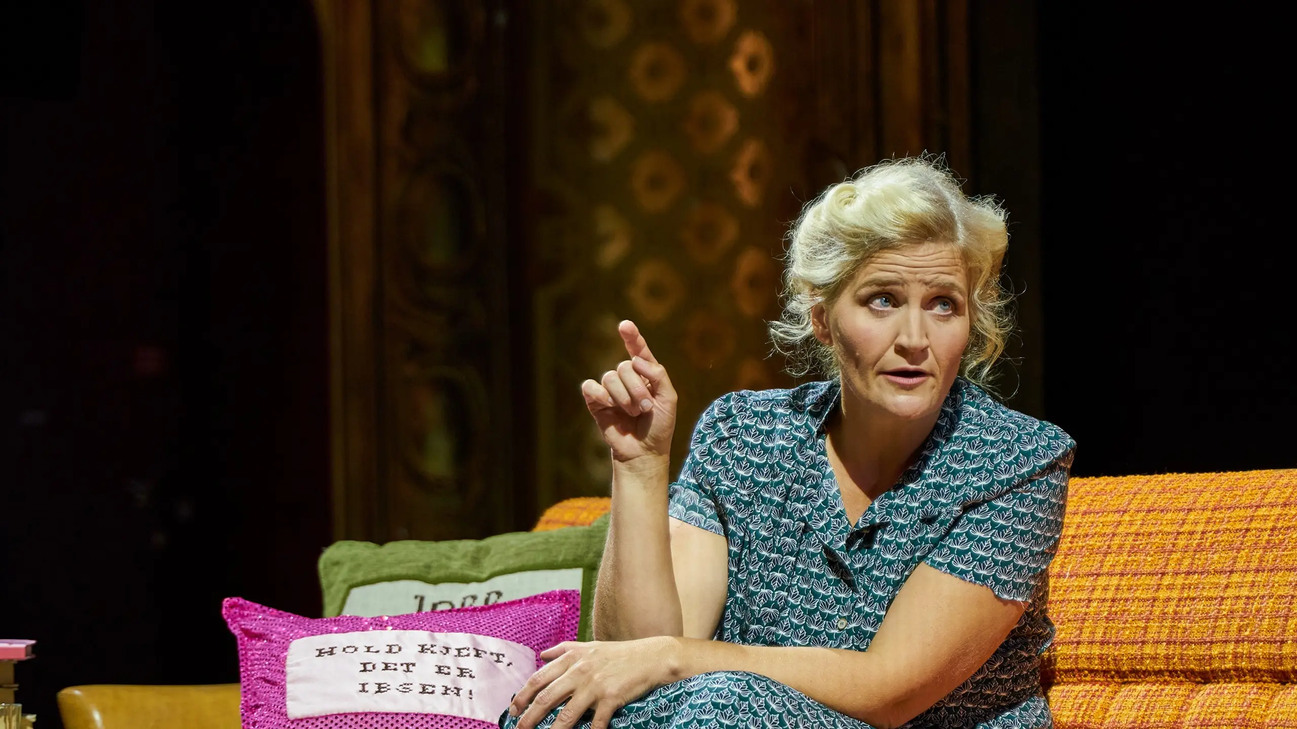 Kvinne sitter i en oransje sofa og gestikulerer, ved siden av henne ligger to puter med teksten "Hold kjeft, det er Ibsen!"