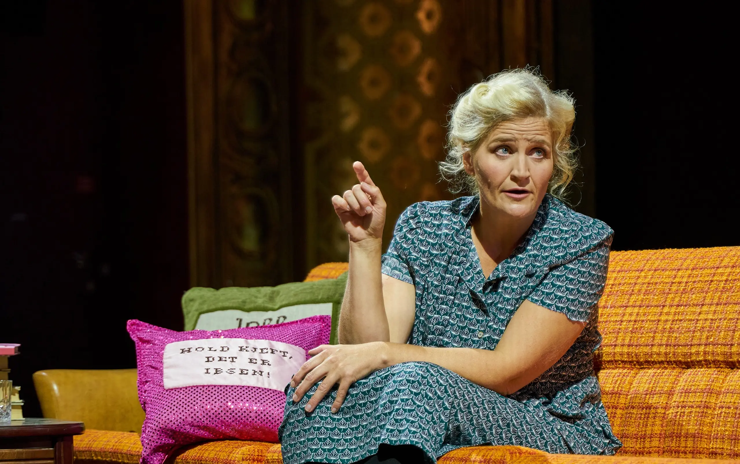 Kvinne sitter i en oransje sofa og gestikulerer, ved siden av henne ligger to puter med teksten "Hold kjeft, det er Ibsen!"