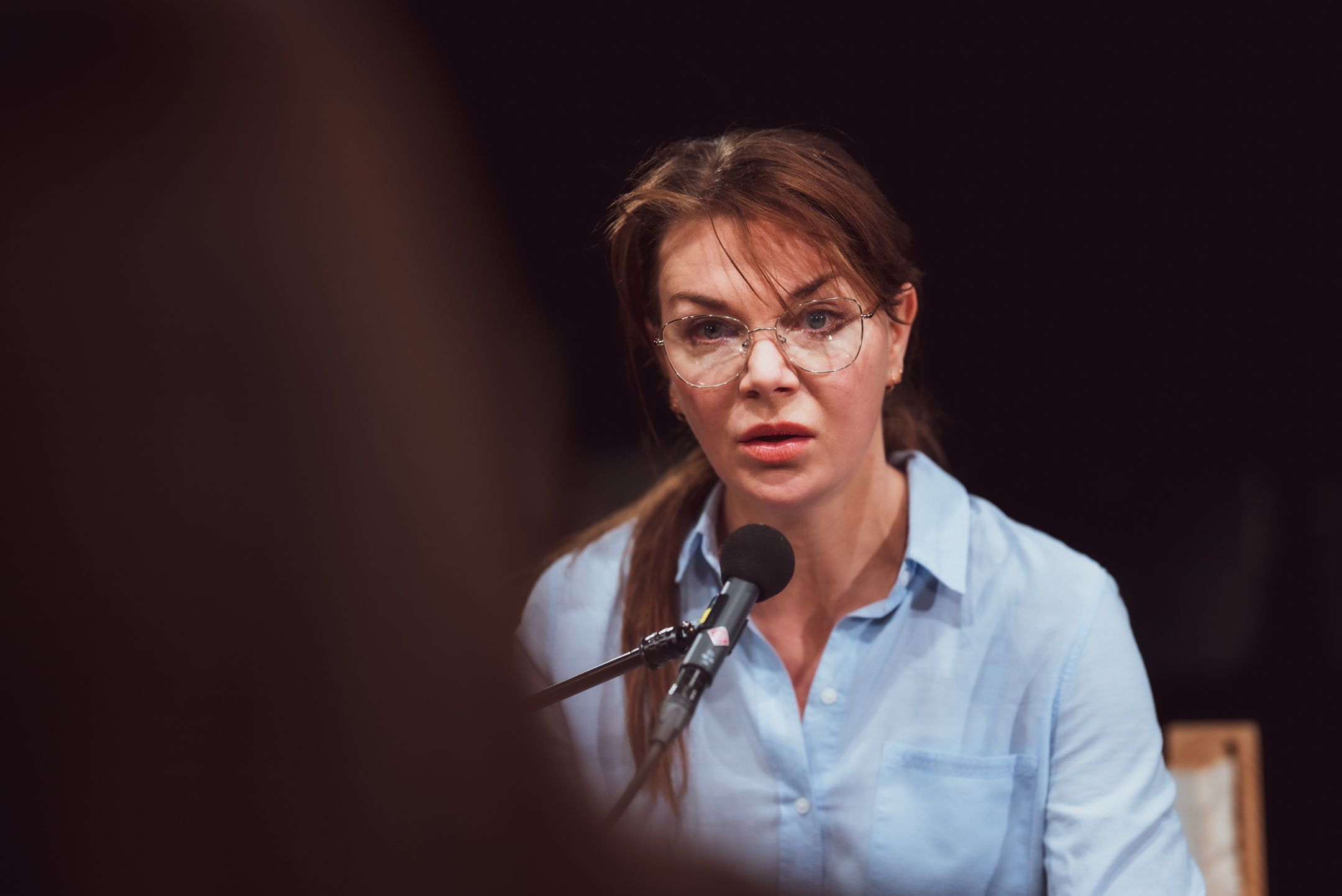 Person med brunt hår og briller snakker i mikrofon med alvorlig uttrykk, kledd i lyseblå skjorte.