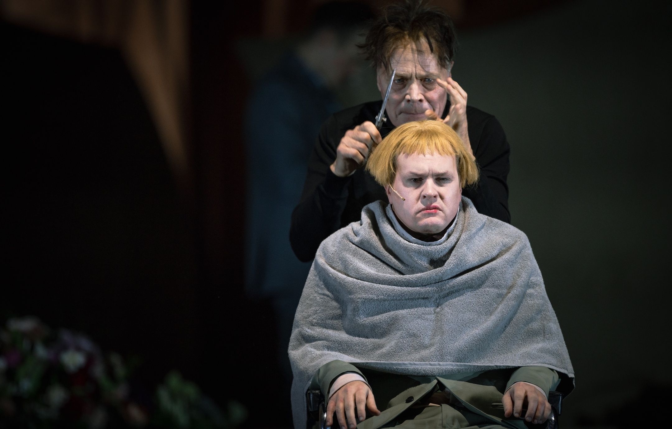 Person i stol med blond parykk får håret klippet av en annen person i sort, begge med alvorlig uttrykk.