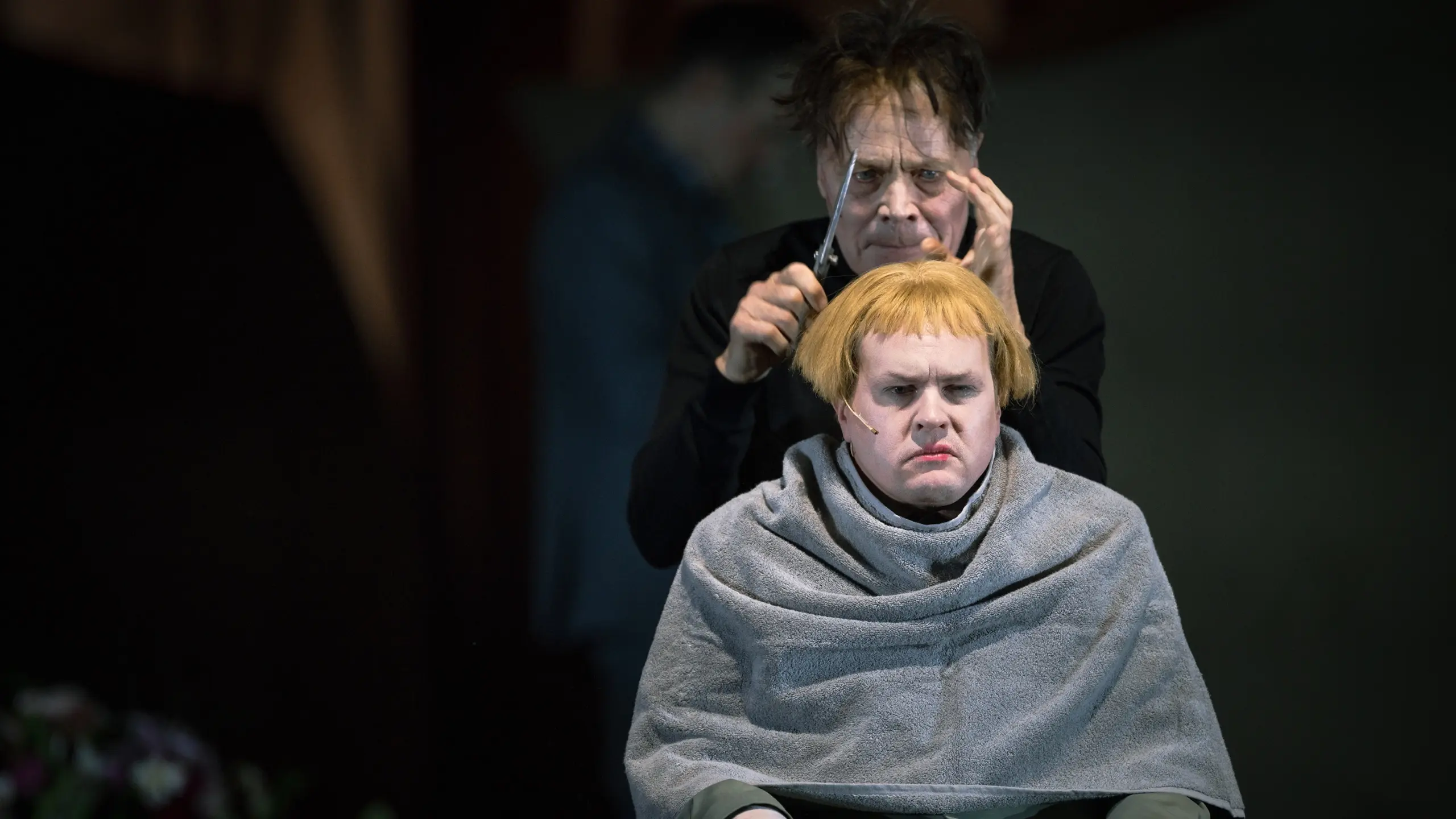 Person i stol med blond parykk får håret klippet av en annen person i sort, begge med alvorlig uttrykk.