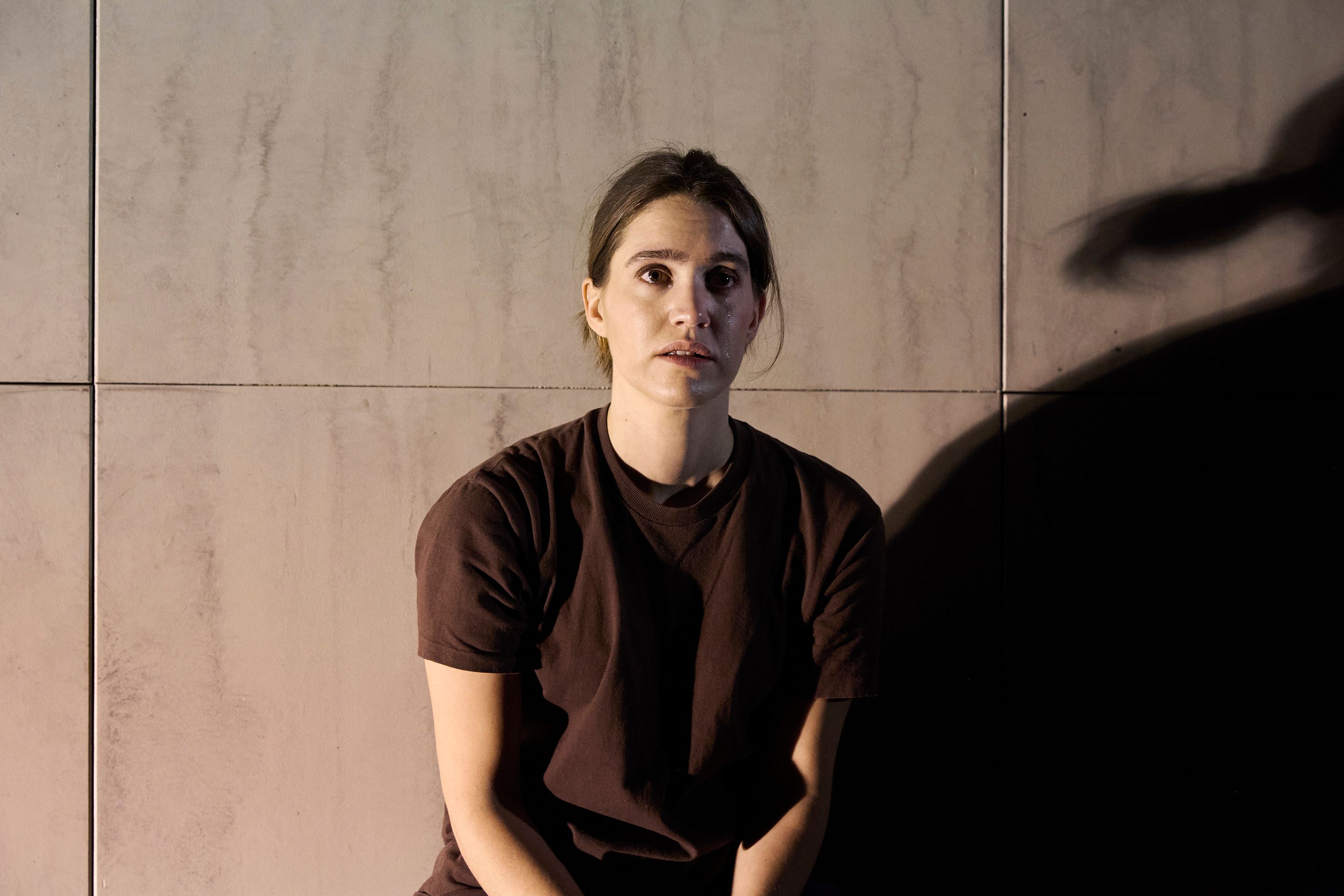 En kvinne i brun t-skjorte sitter mot en grå vegg, ser rett frem med alvorlig uttrykk i dempet belysning. En tåre renner ned kinnet.