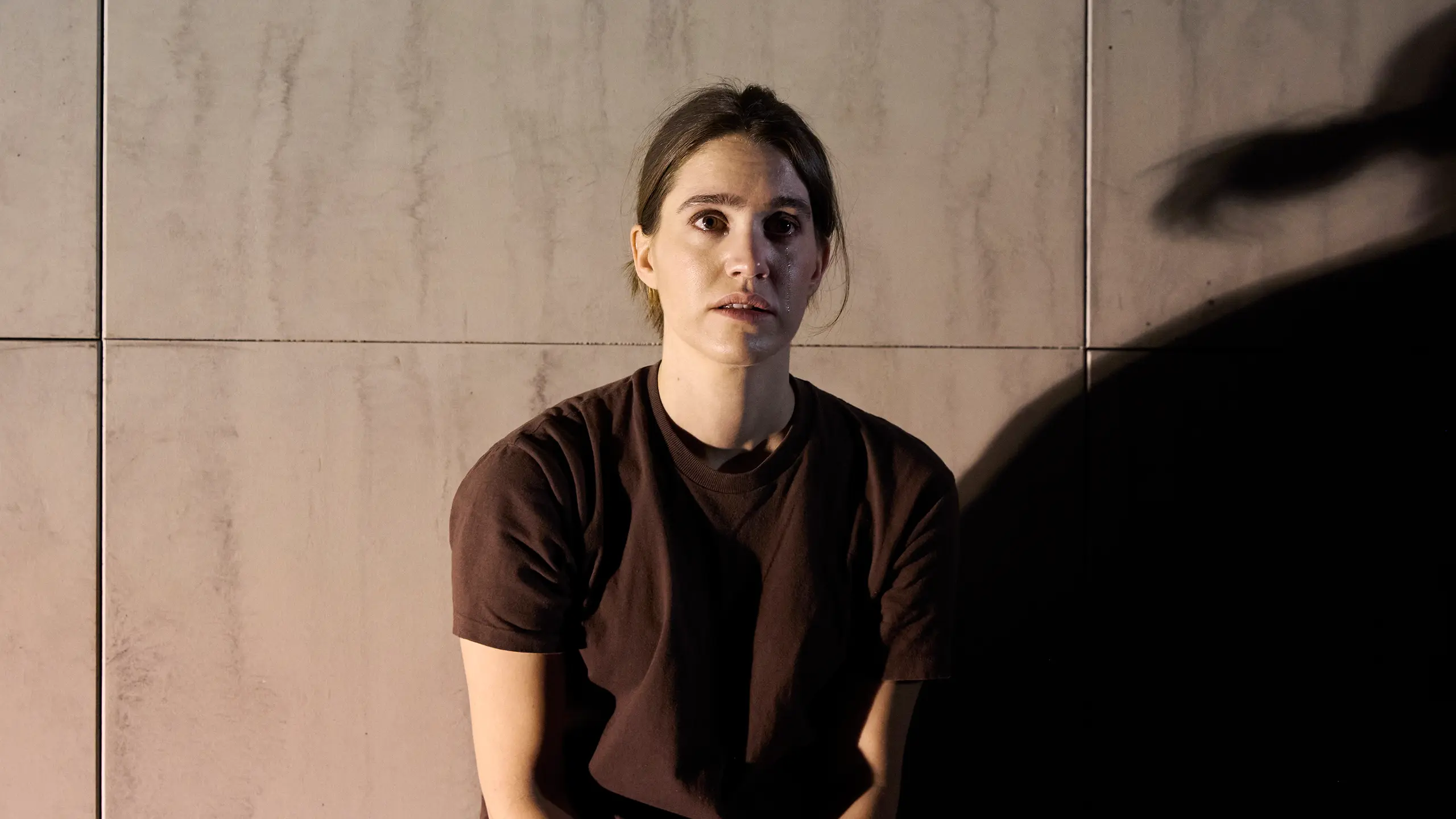 En kvinne i brun t-skjorte sitter mot en grå vegg, ser rett frem med alvorlig uttrykk i dempet belysning. En tåre renner ned kinnet.