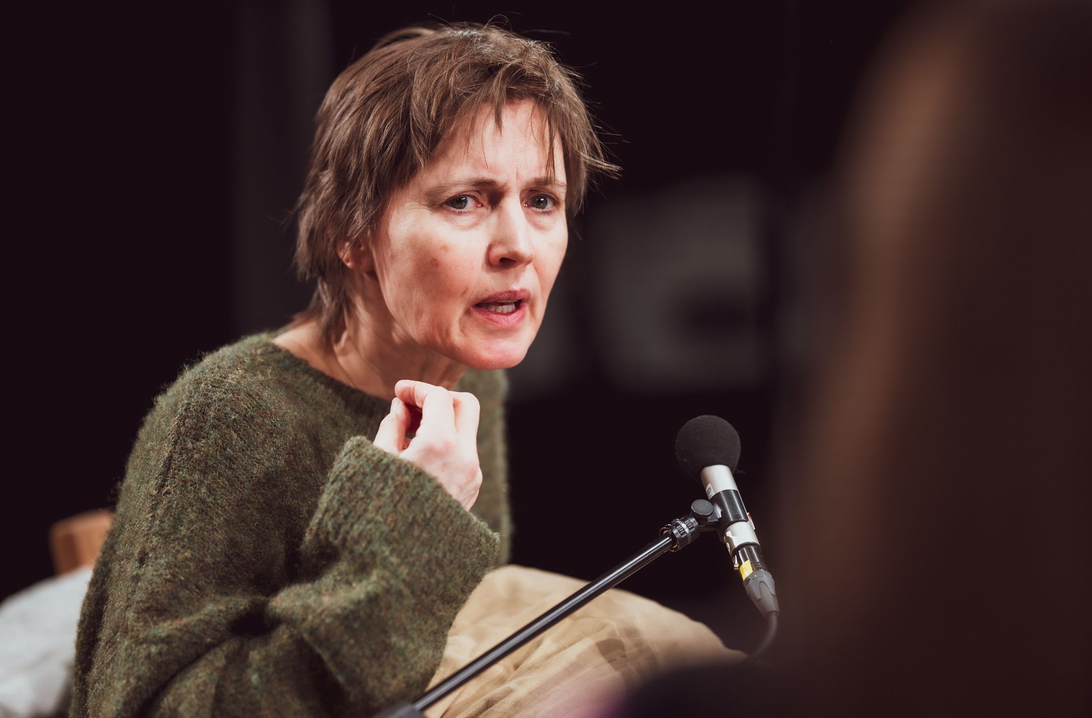 Person med kort brunt hår i grønn strikkegenser snakker inn i mikrofon med alvorlig uttrykk og løftet hånd.