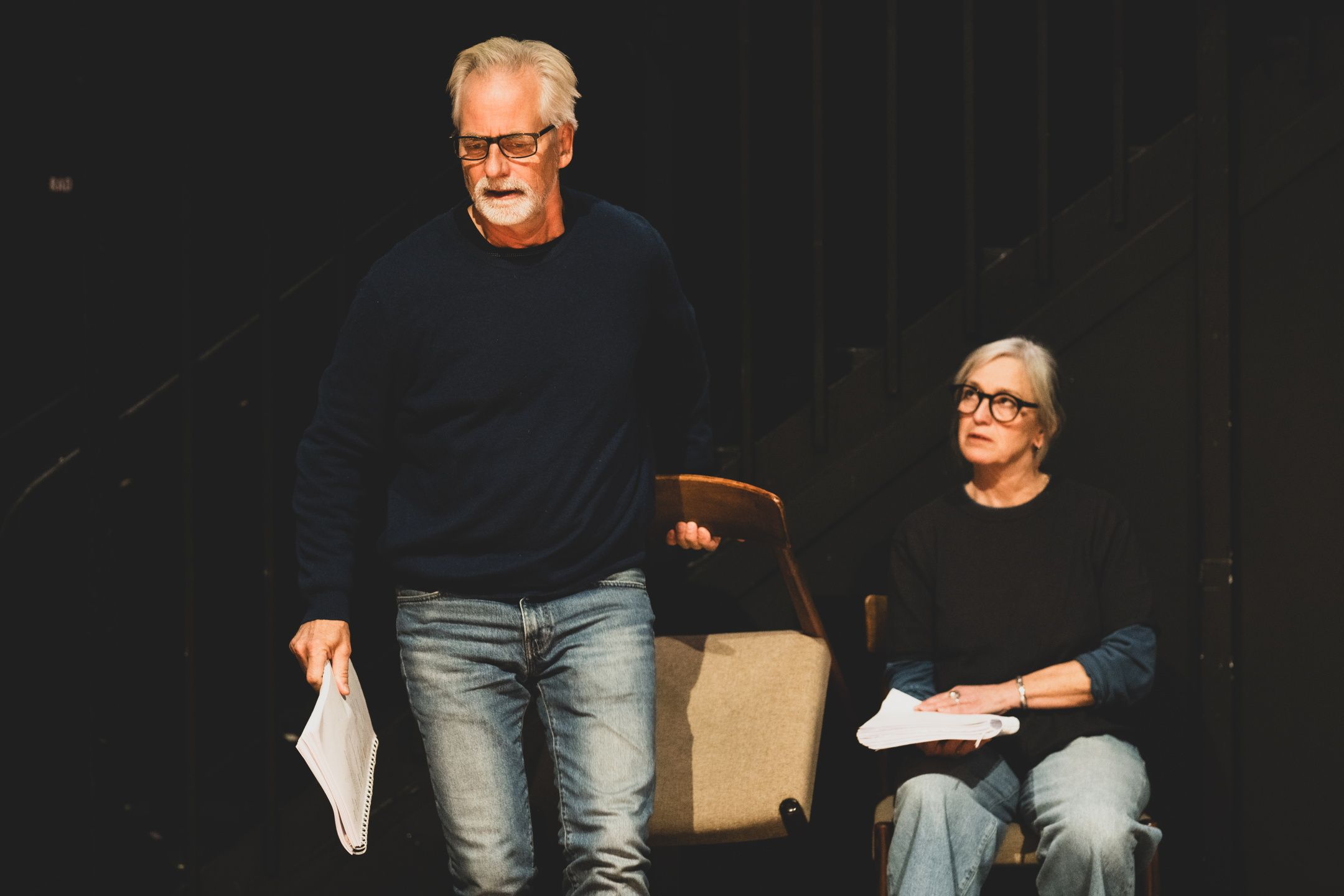 Mann som løfter en stol og holder manus med den andre hånda, mens en kvinne sitter på en stol med manus ved siden av