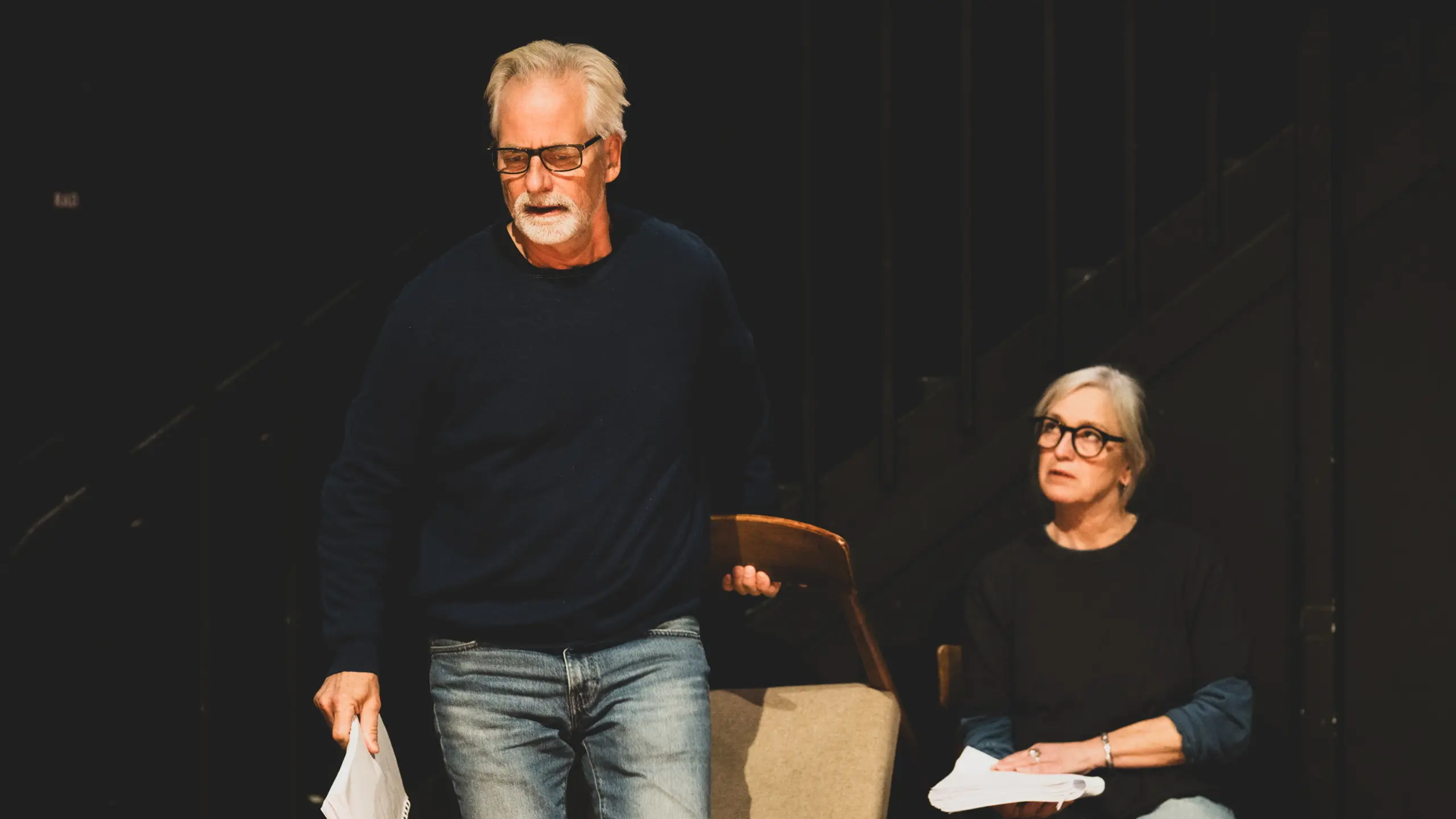 Mann som løfter en stol og holder manus med den andre hånda, mens en kvinne sitter på en stol med manus ved siden av