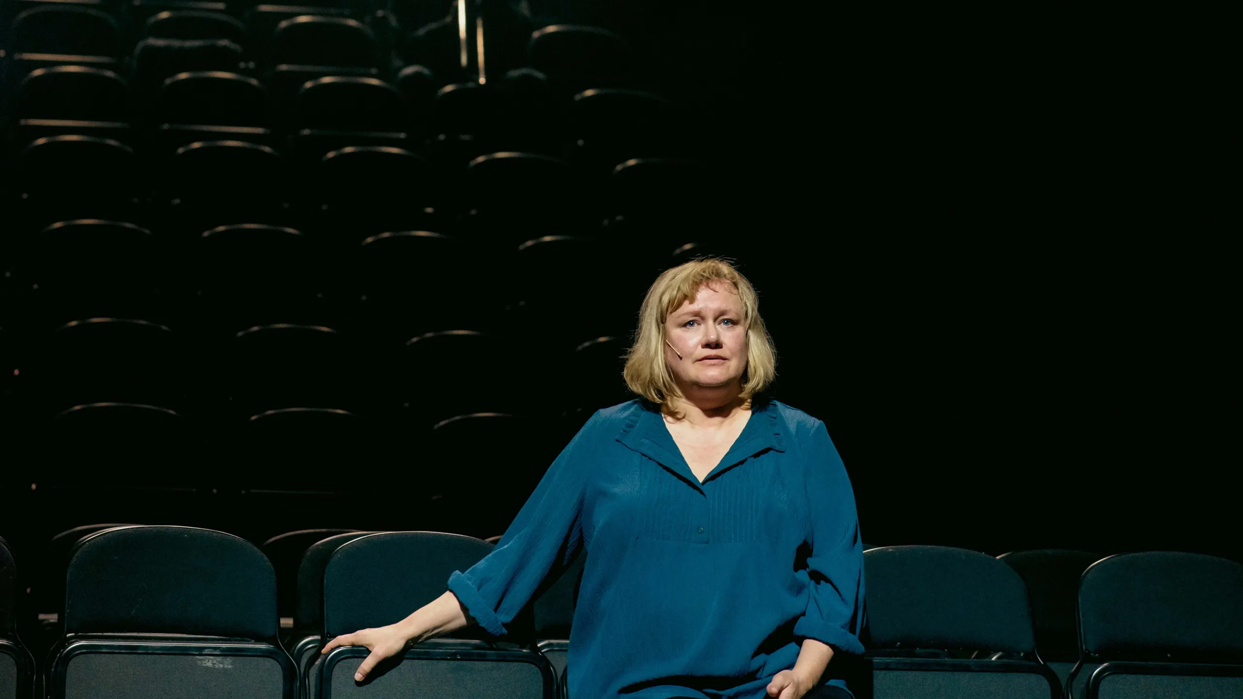 Kvinne sitter fremmerst i et mørkt parti. Bak skimter vi en mann med trombone