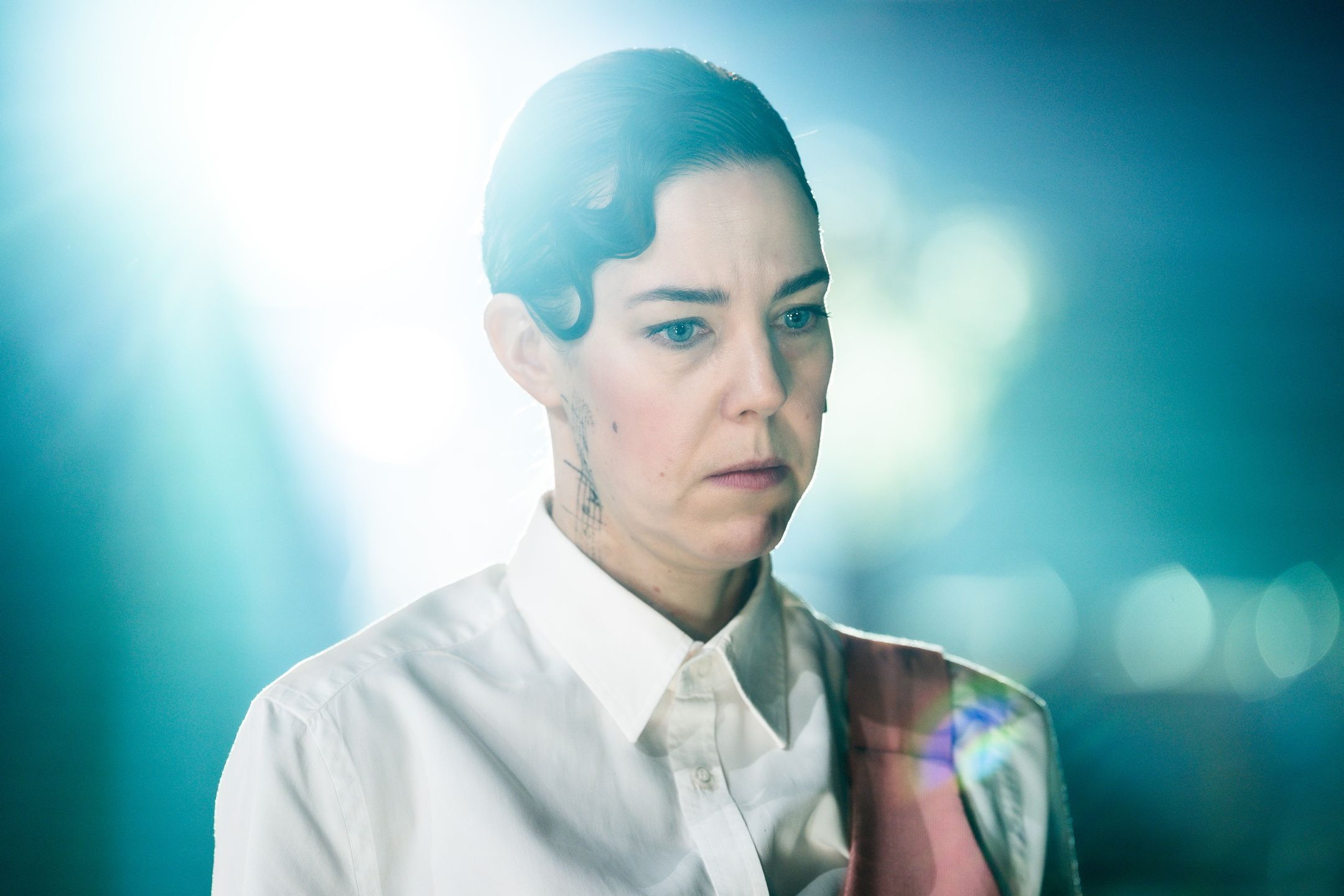 En kvinnelig skuespiller i hvit skjorte og rosa element ser alvorlig og ettertenksom ut. Hun har påtegnet grafiske mønstre på halsen. Et sterkt lys skinner inn fra venstre og skaper en dramatisk og filmatisk effekt.