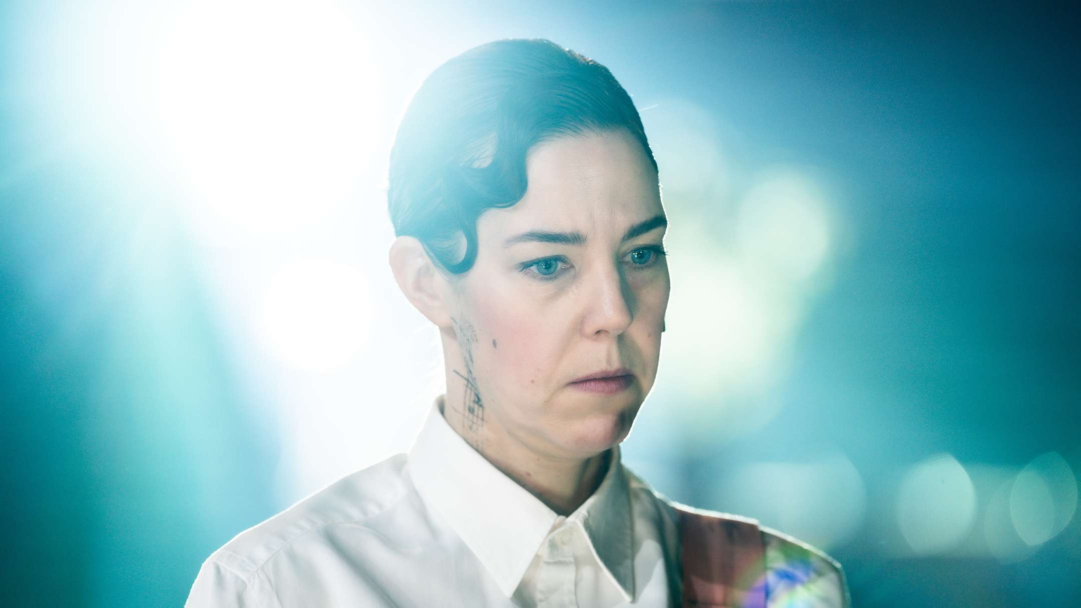 En kvinnelig skuespiller i hvit skjorte og rosa element ser alvorlig og ettertenksom ut. Hun har påtegnet grafiske mønstre på halsen. Et sterkt lys skinner inn fra venstre og skaper en dramatisk og filmatisk effekt.