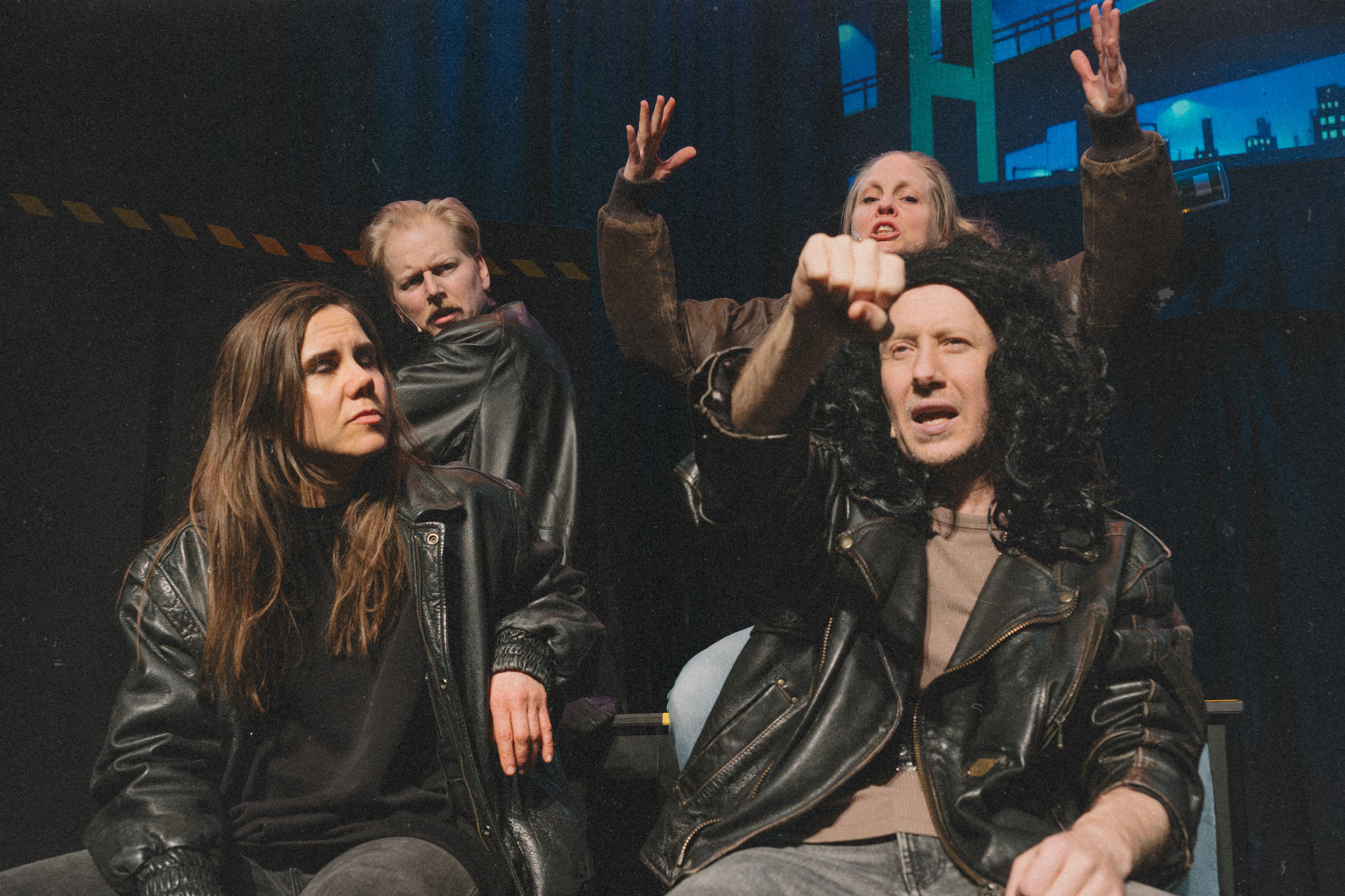 Fire personer i skinnjakker sitter og gestikulerer dramatisk, én peker fremover med knyttet neve