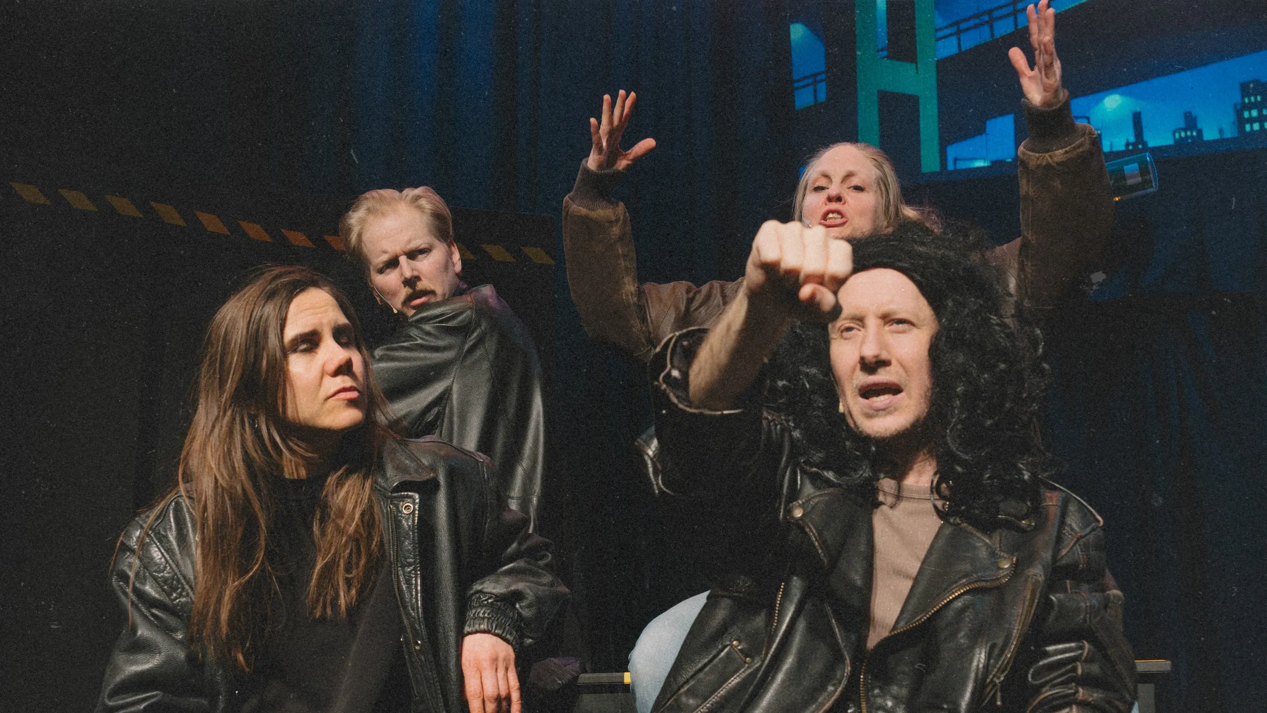 Fire personer i skinnjakker sitter og gestikulerer dramatisk, én peker fremover med knyttet neve