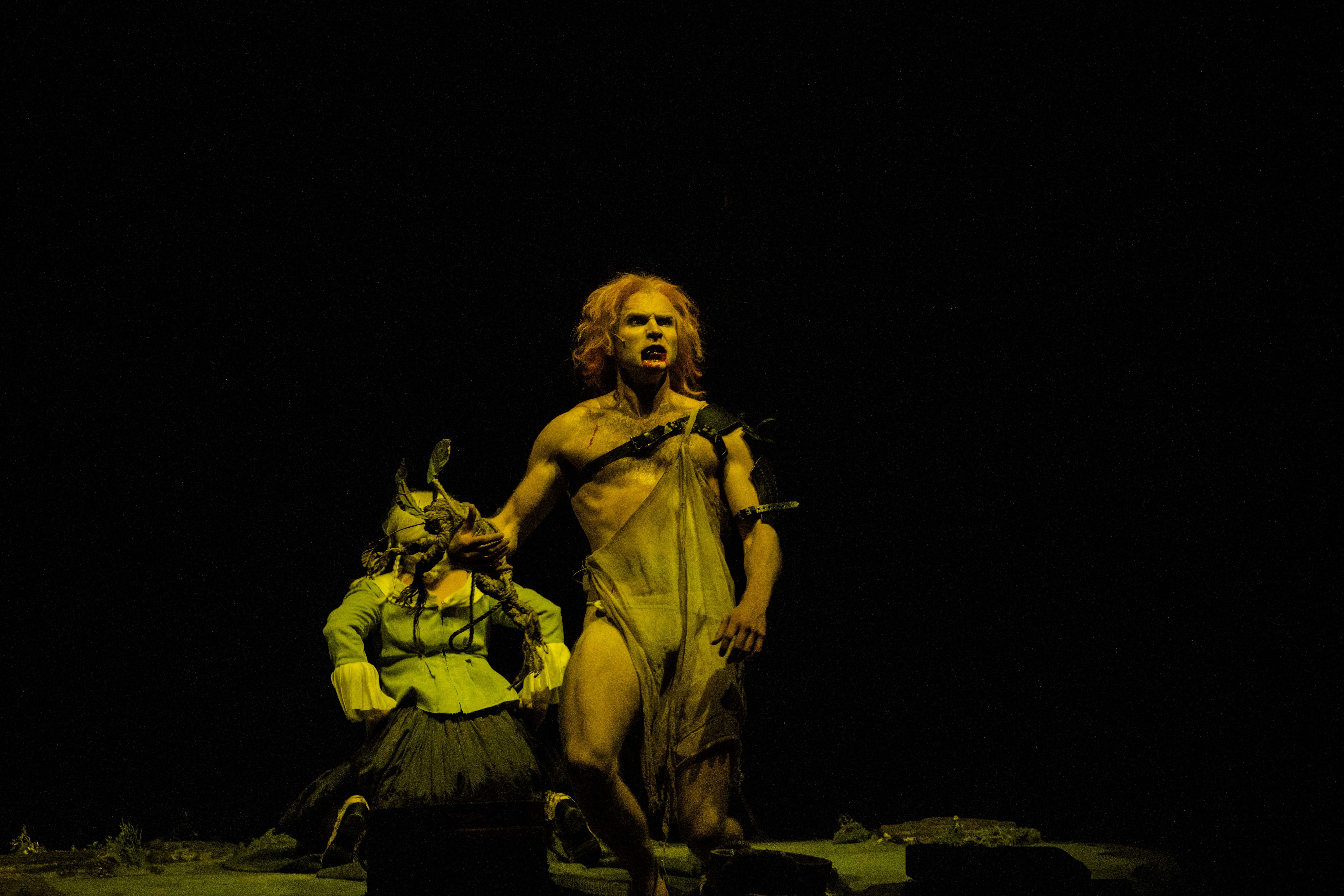 En barbrystet mann med rødt hår kneler ved siden av en sittende figur i grønt kostyme, dramatisk lyssatt mot mørk bakgrunn.