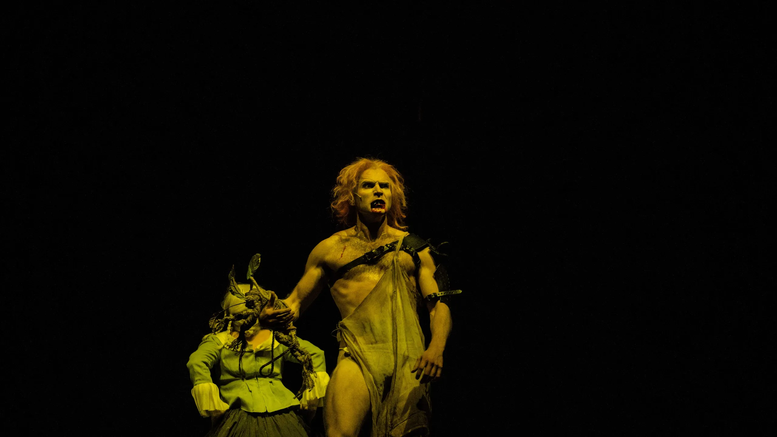 En barbrystet mann med rødt hår kneler ved siden av en sittende figur i grønt kostyme, dramatisk lyssatt mot mørk bakgrunn.