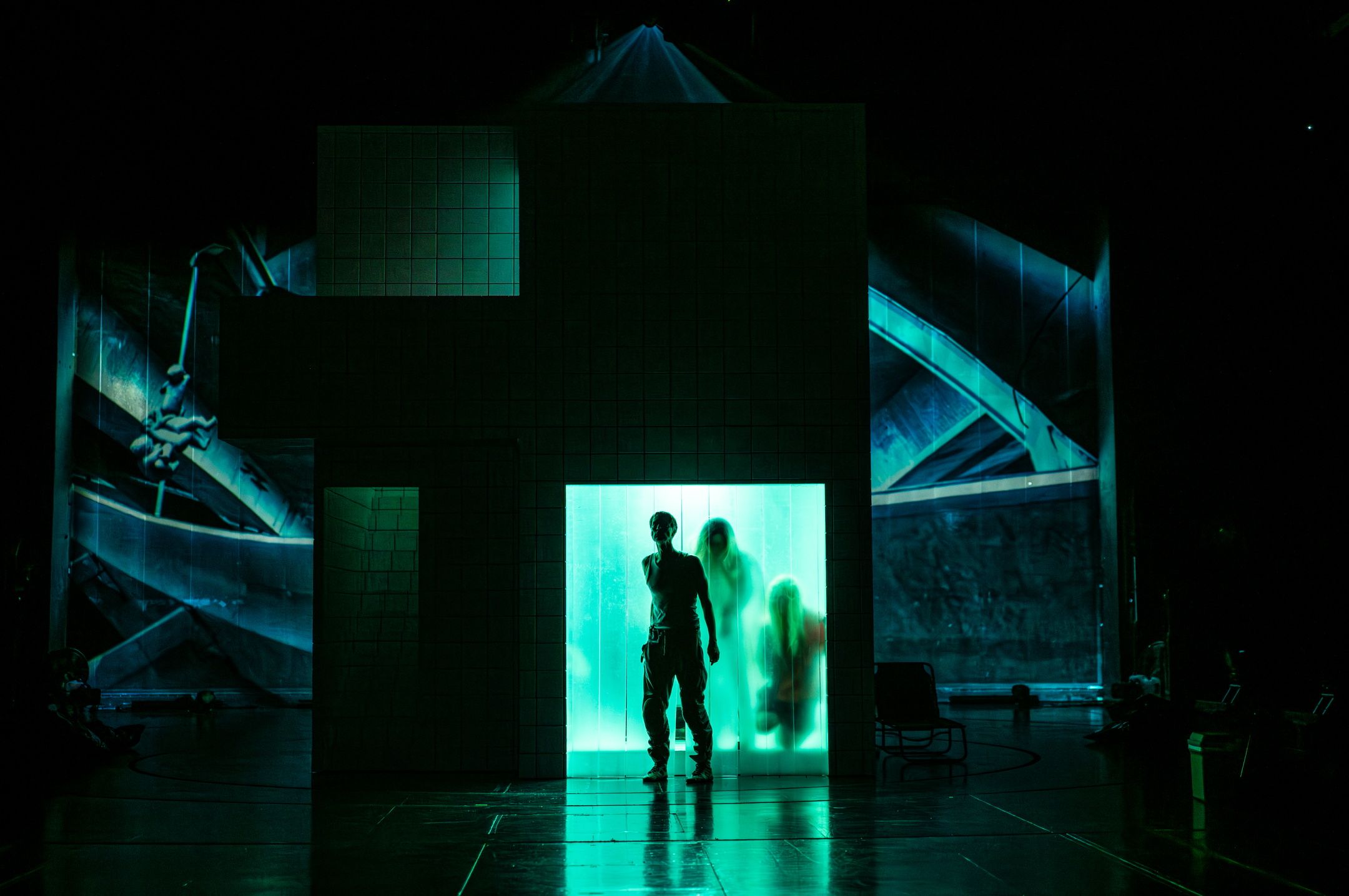 Mørklagt scene med grønn kubeformet struktur, tre skikkelser bak opplyst plastvegg