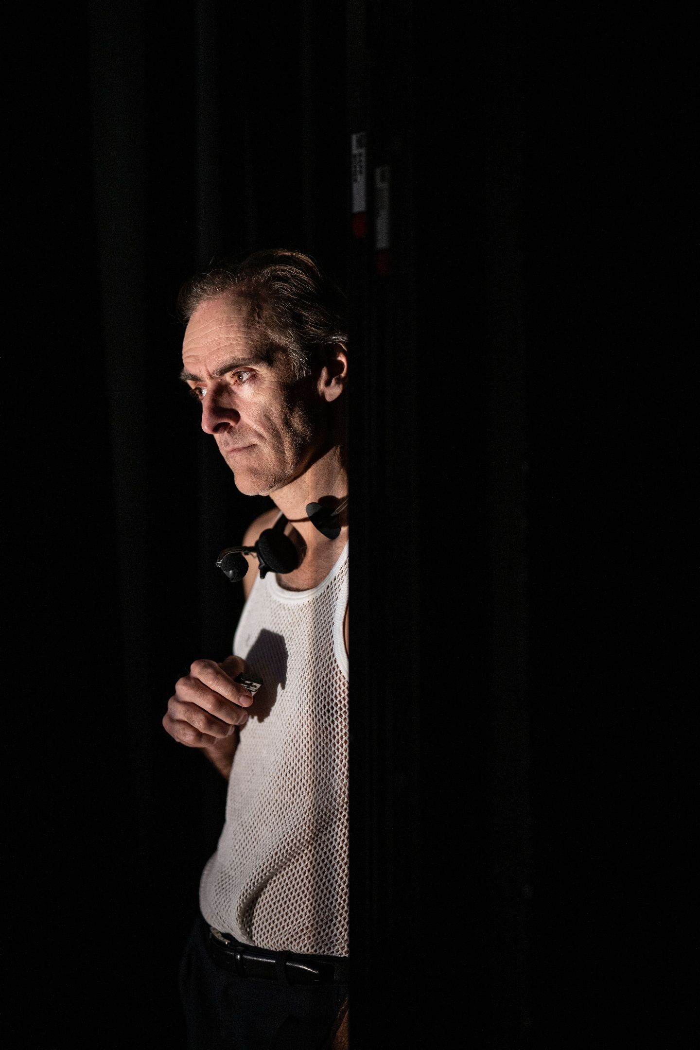 Mann i hvit nettingtrøye og hodetelefoner ser alvorlig ut bak et sceneteppe