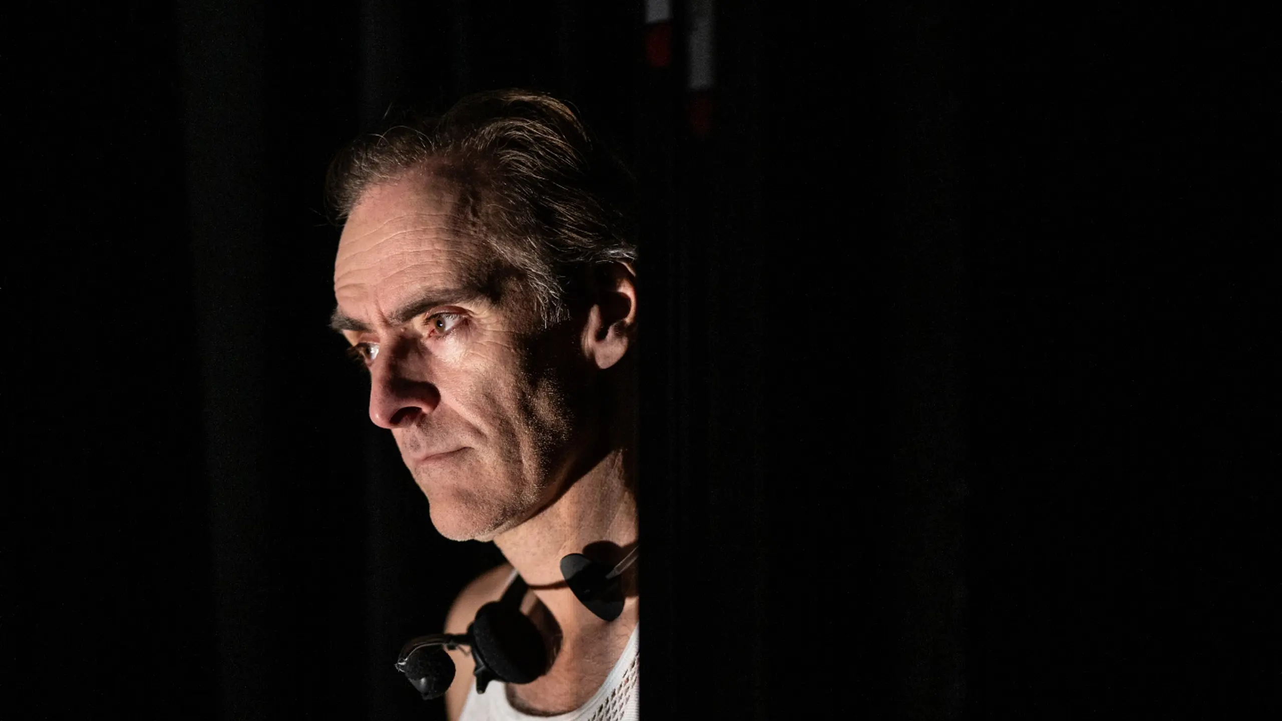 Mann i hvit nettingtrøye og hodetelefoner ser alvorlig ut bak et sceneteppe