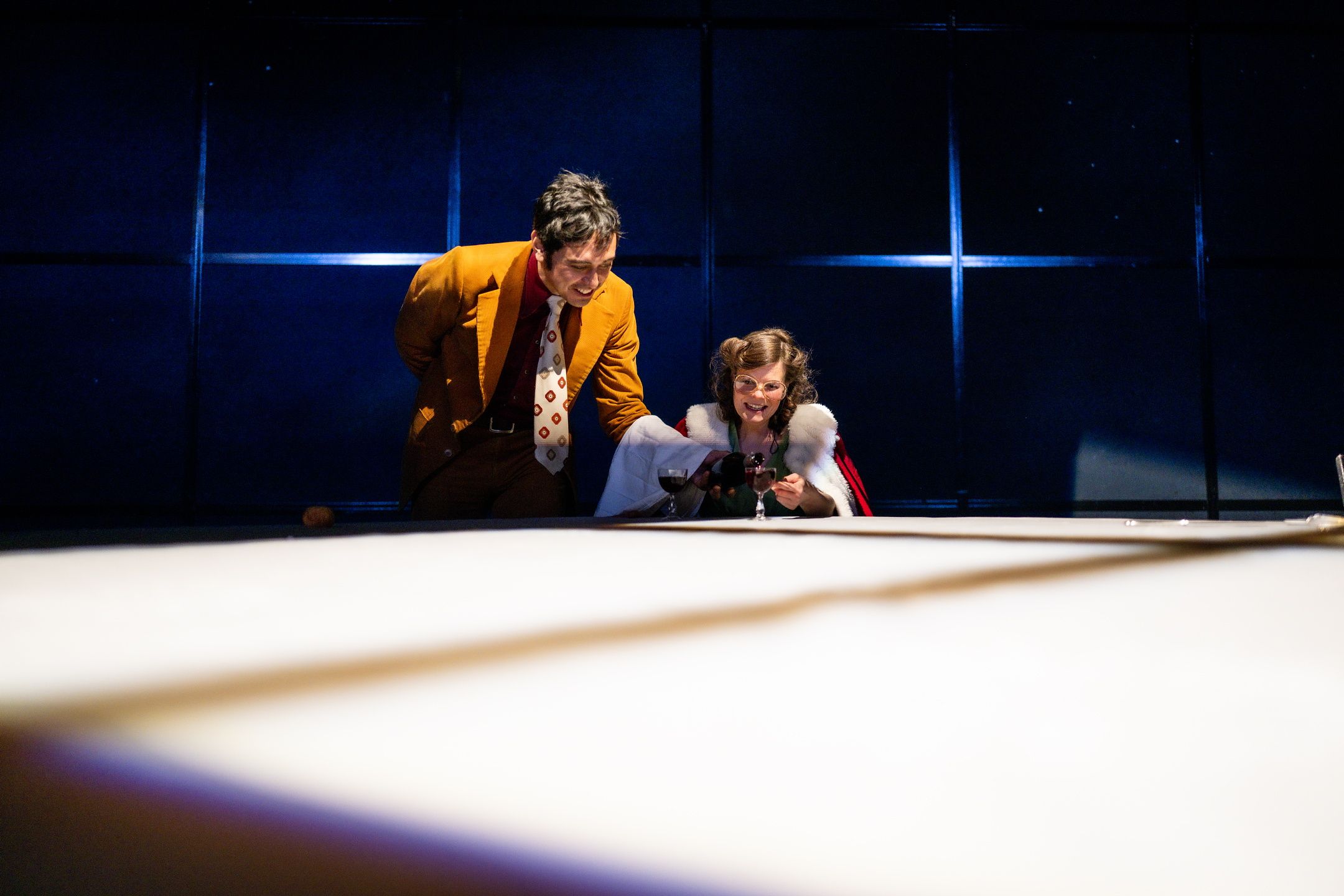 Mann i orange dress står smilende ved siden av en smilende kvinne vmed nissekappe. Han skjenker henne et glass rødvin