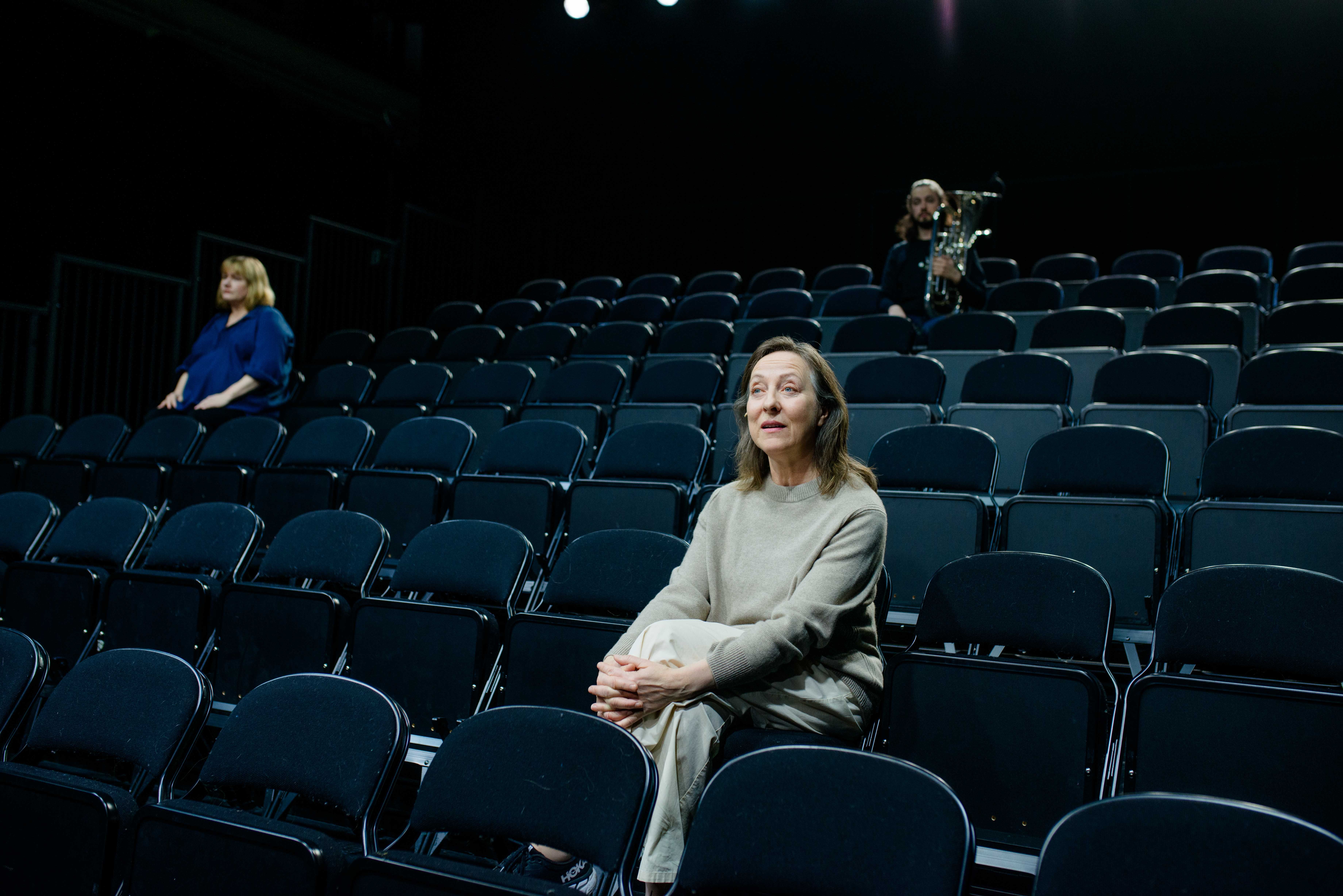 To kvinner sitter i et tomt mørkt amfi. Bak ser vi en mann med baryton