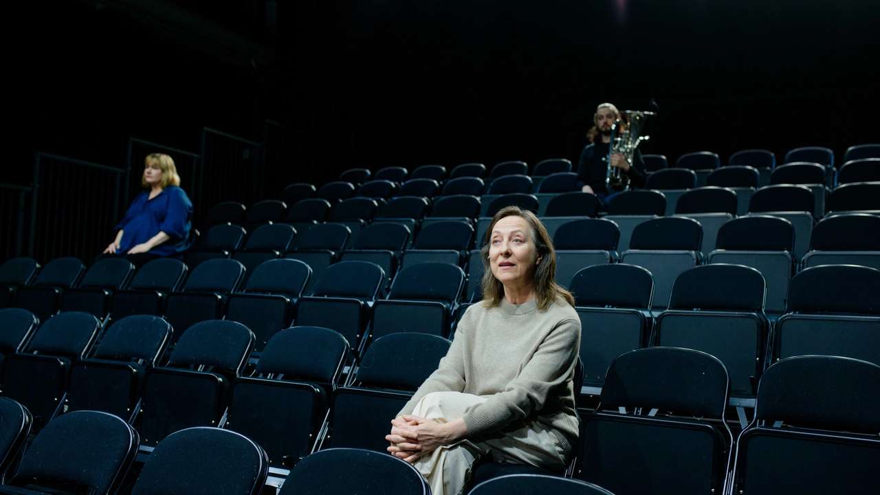 To kvinner sitter i et tomt mørkt amfi. Bak ser vi en mann med baryton