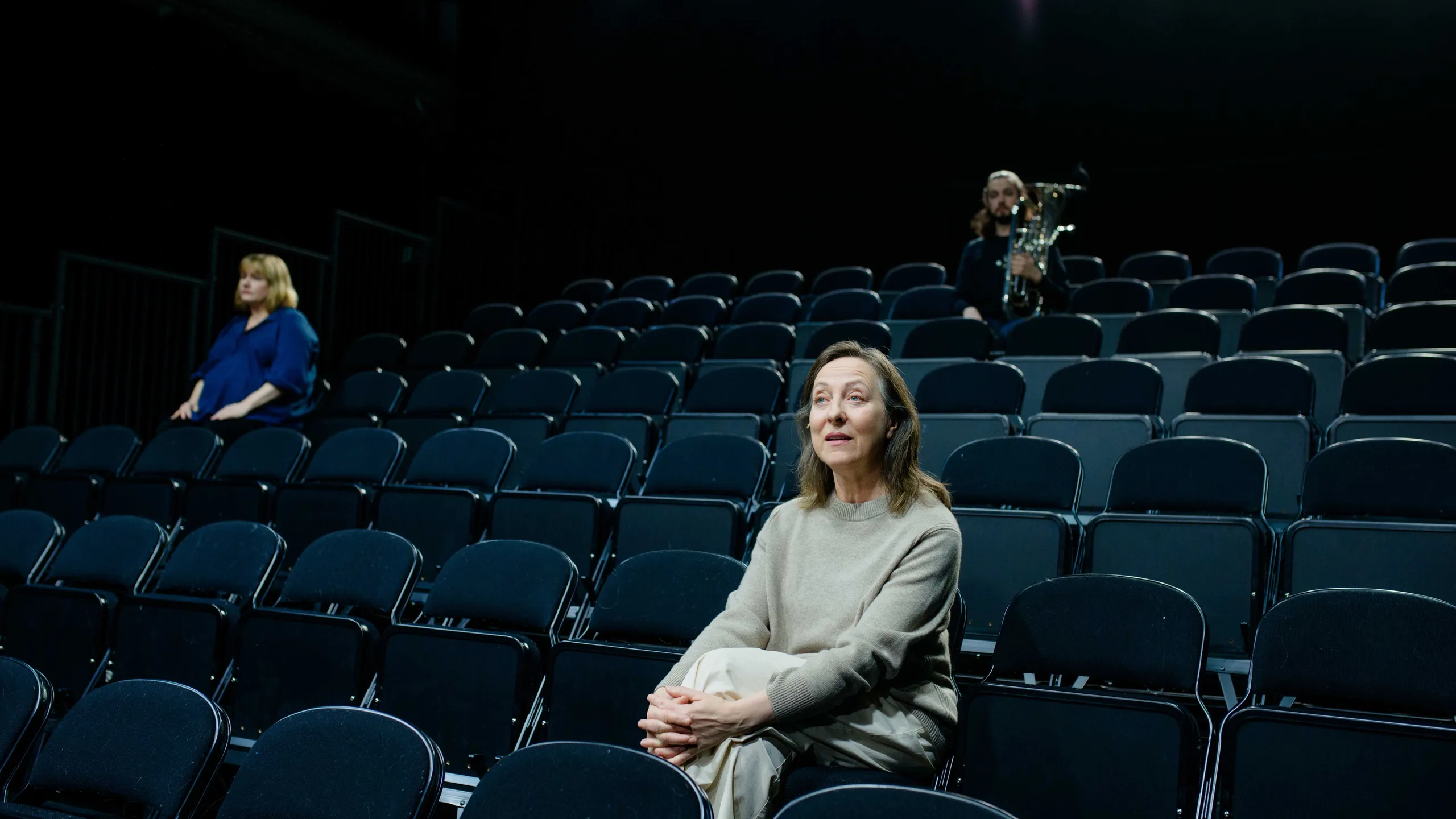 To kvinner sitter i et tomt mørkt amfi. Bak ser vi en mann med baryton