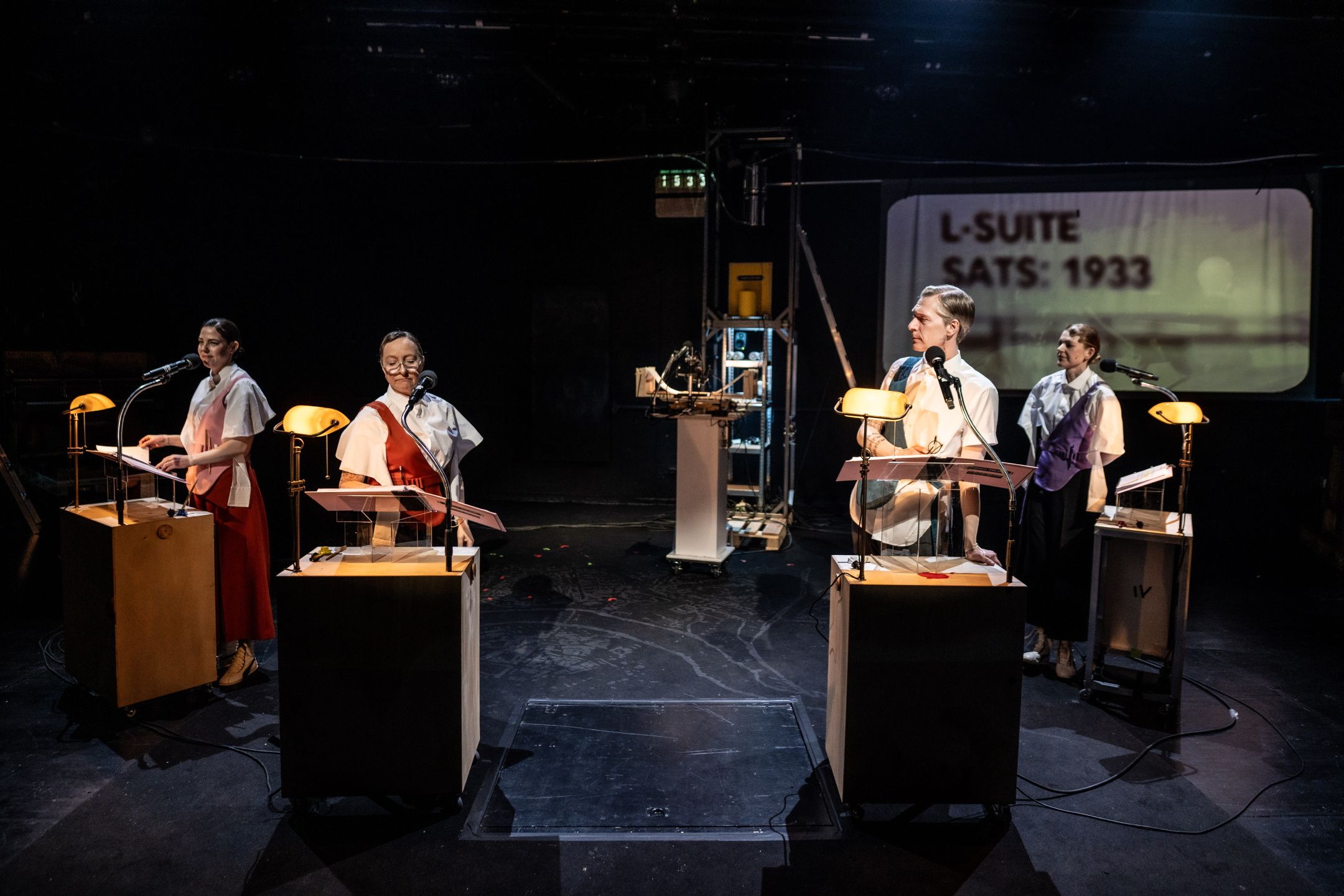 Fire skuespillere står ved hver sin pult med mikrofon og små lamper. Bak dem ser vi projisert tekst: “L-SUITE / SATS: 1933”.