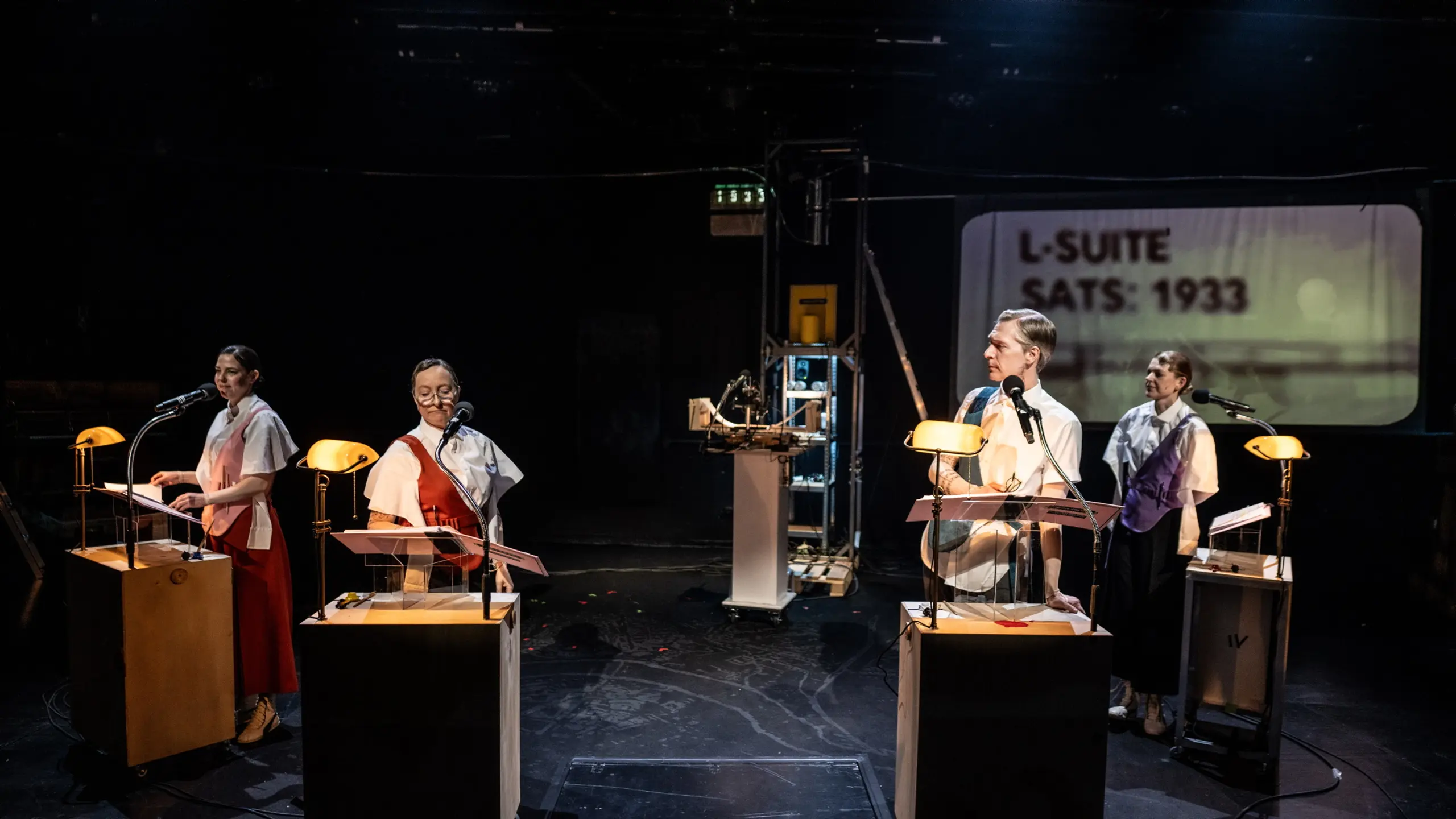 Fire skuespillere står ved hver sin pult med mikrofon og små lamper. Bak dem ser vi projisert tekst: “L-SUITE / SATS: 1933”.