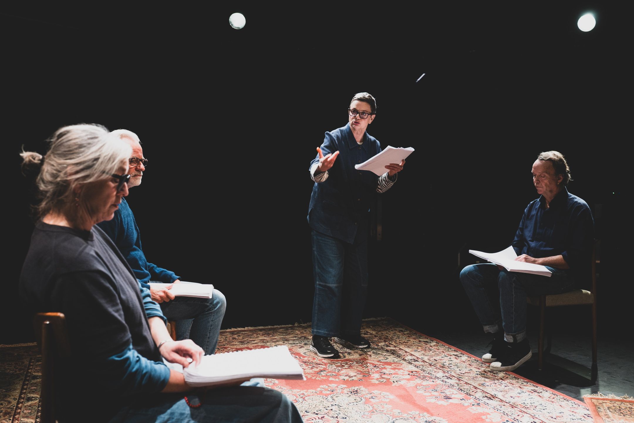 En kvinne står med manus og gestikulerer til to menn og en kvinne som sitter rundt henne med manus