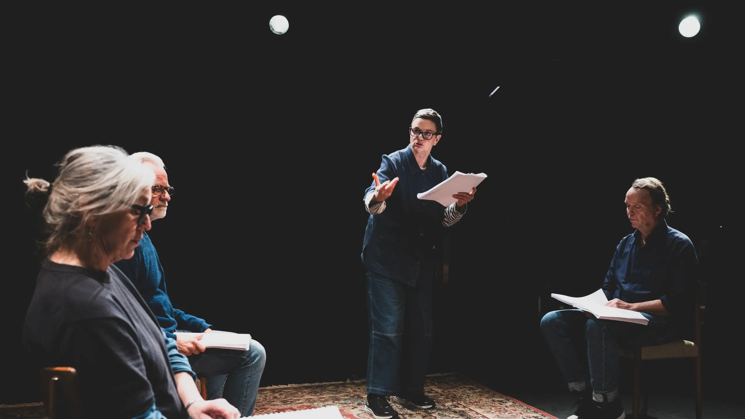 En kvinne står med manus og gestikulerer til to menn og en kvinne som sitter rundt henne med manus