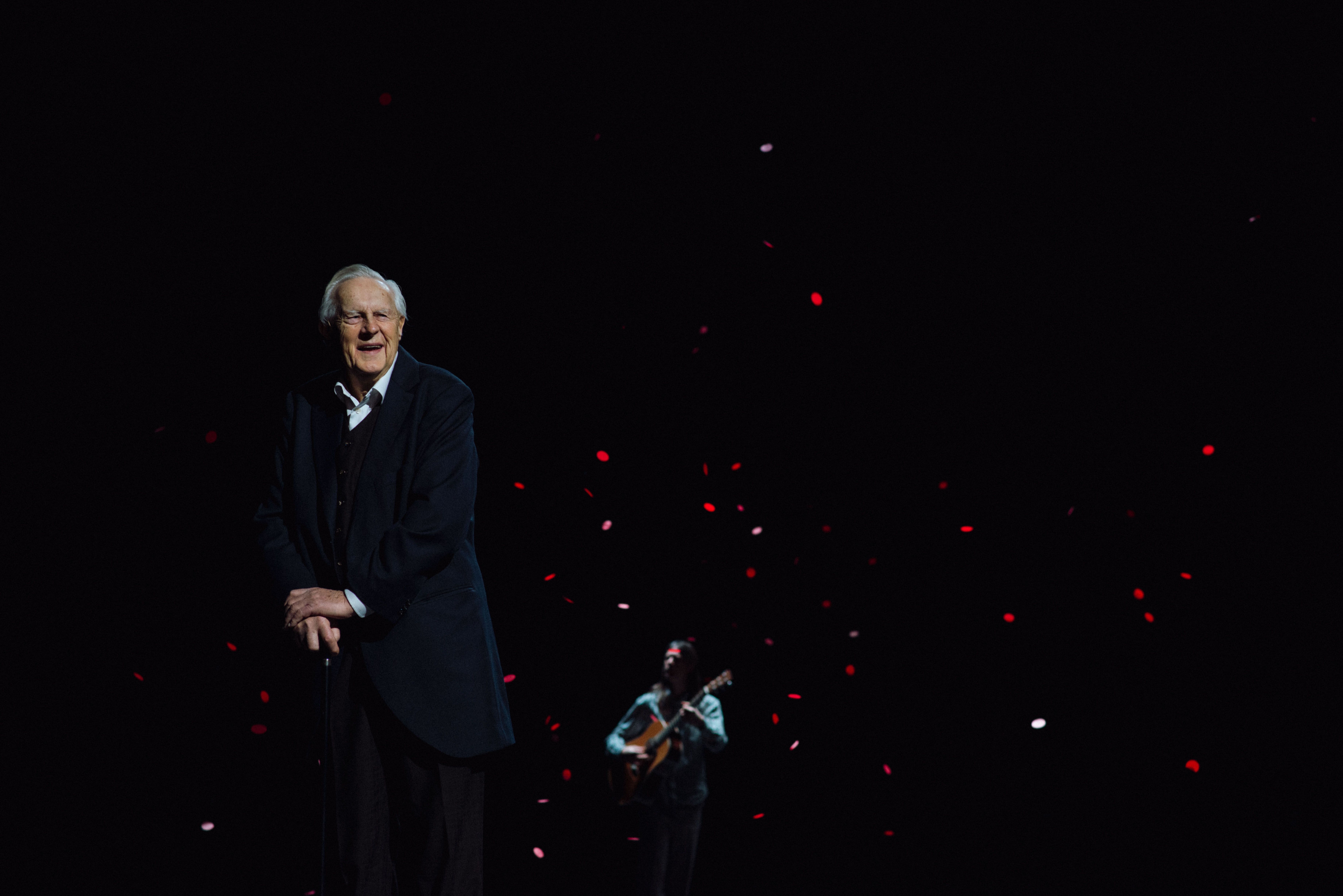 Skuespiller Sverre Anker Ousdal står i sort frakk i mørke omgivelser på scenen. I luften faller det noe som kan ligne på roseblader, og bak ham står det en mann og spiller gitar.