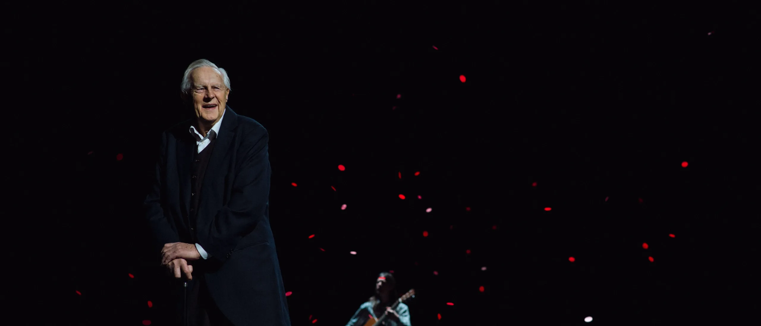 Skuespiller Sverre Anker Ousdal står i sort frakk i mørke omgivelser på scenen. I luften faller det noe som kan ligne på roseblader, og bak ham står det en mann og spiller gitar.
