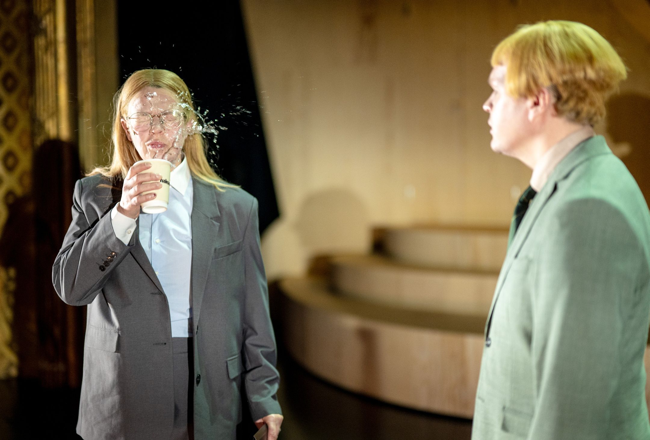 Væske spruter ut av en kopp i ansiktet på person med langt hår og briller, en annen person ser på.