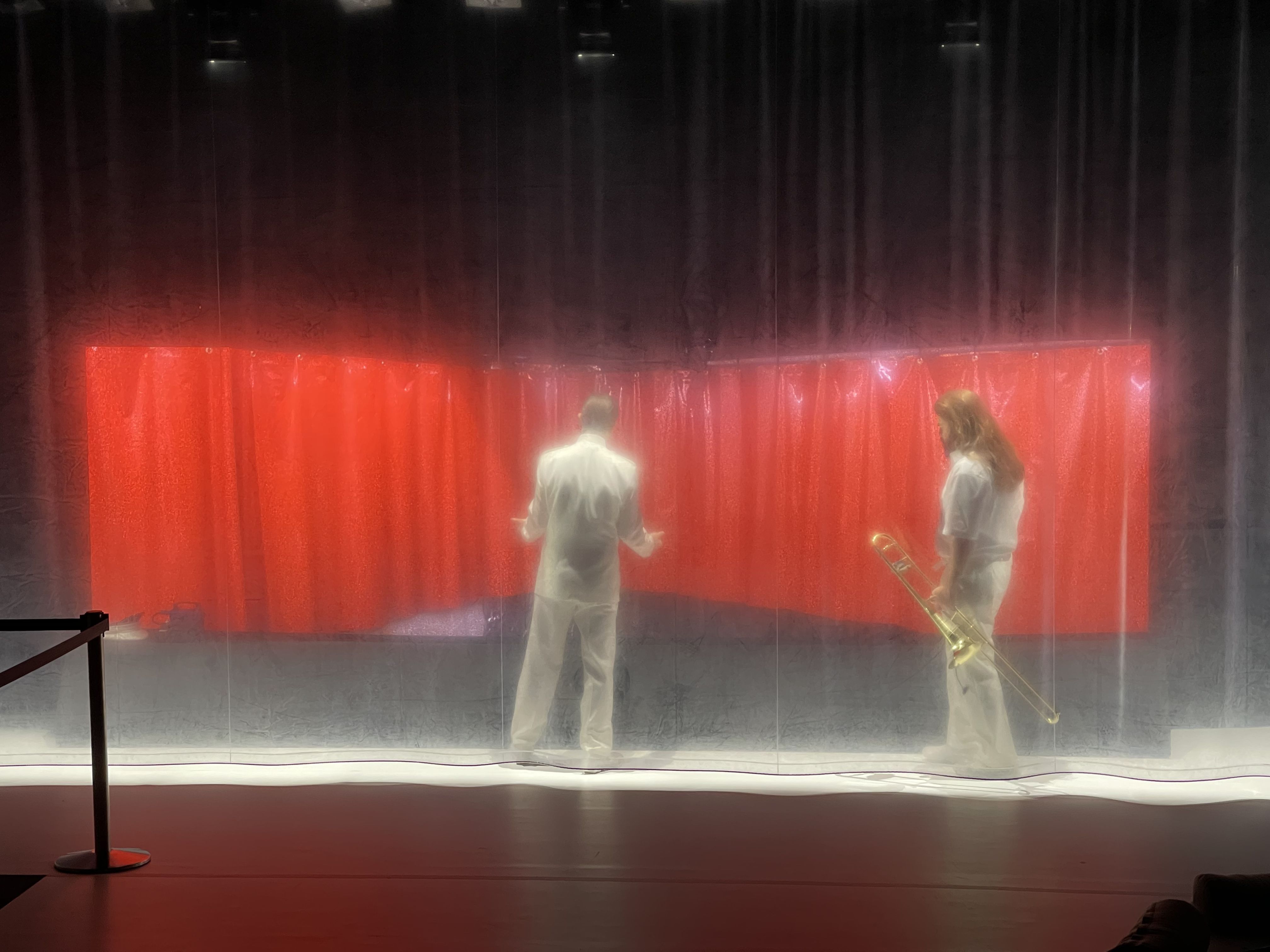 Prøvebilder fra scenen på forestillingen Kallocain - vi ser to skuespillere i hvite klær bak et gjennomsiktig forheng i plast. De står foran noe som er ut som et rødt forheng, og den ene holder en trombone i hånden.